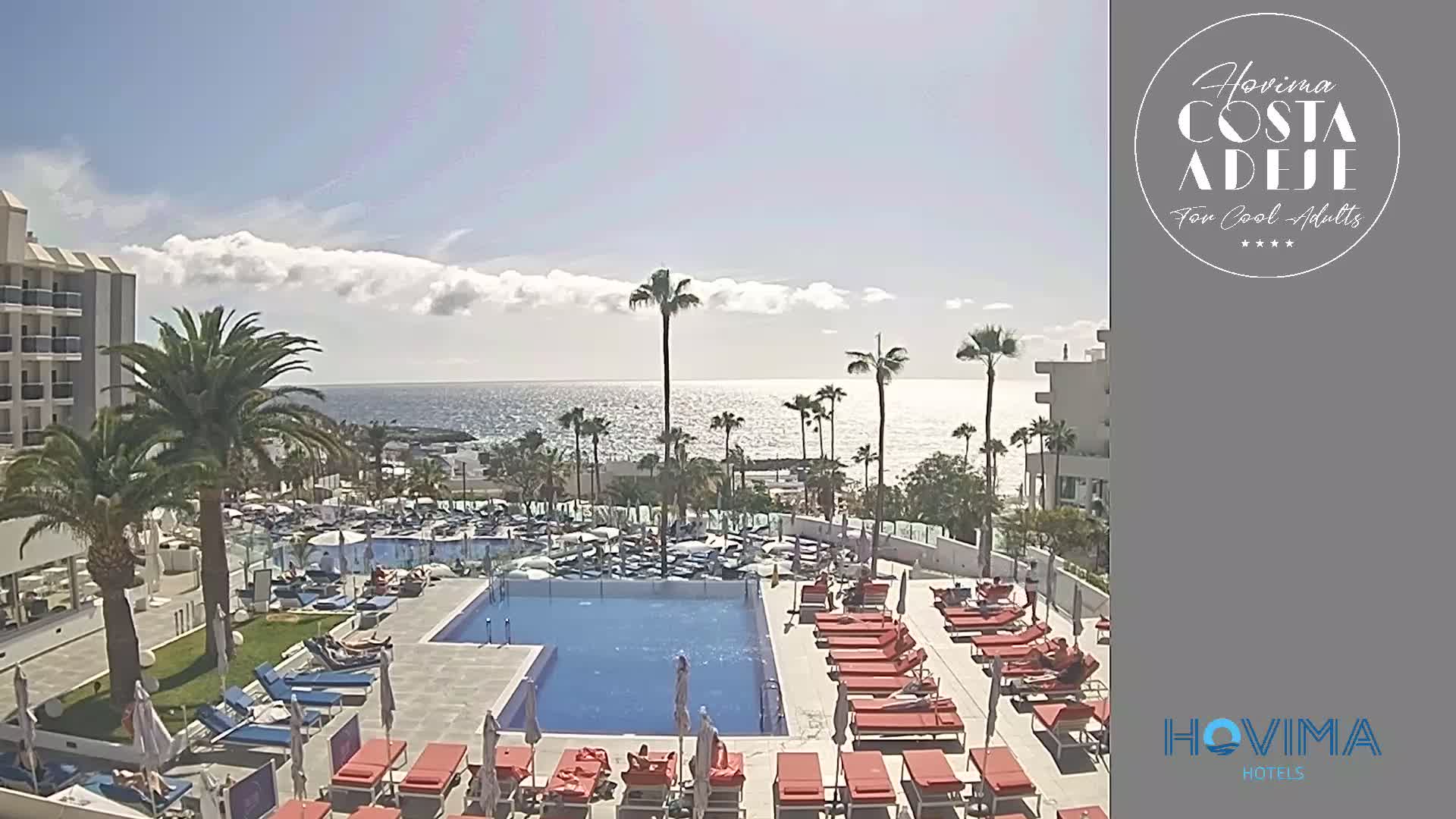 A resort pool area with numerous orange lounge chairs surrounds a large swimming pool, palm trees, and a calm ocean under a partly cloudy sky.