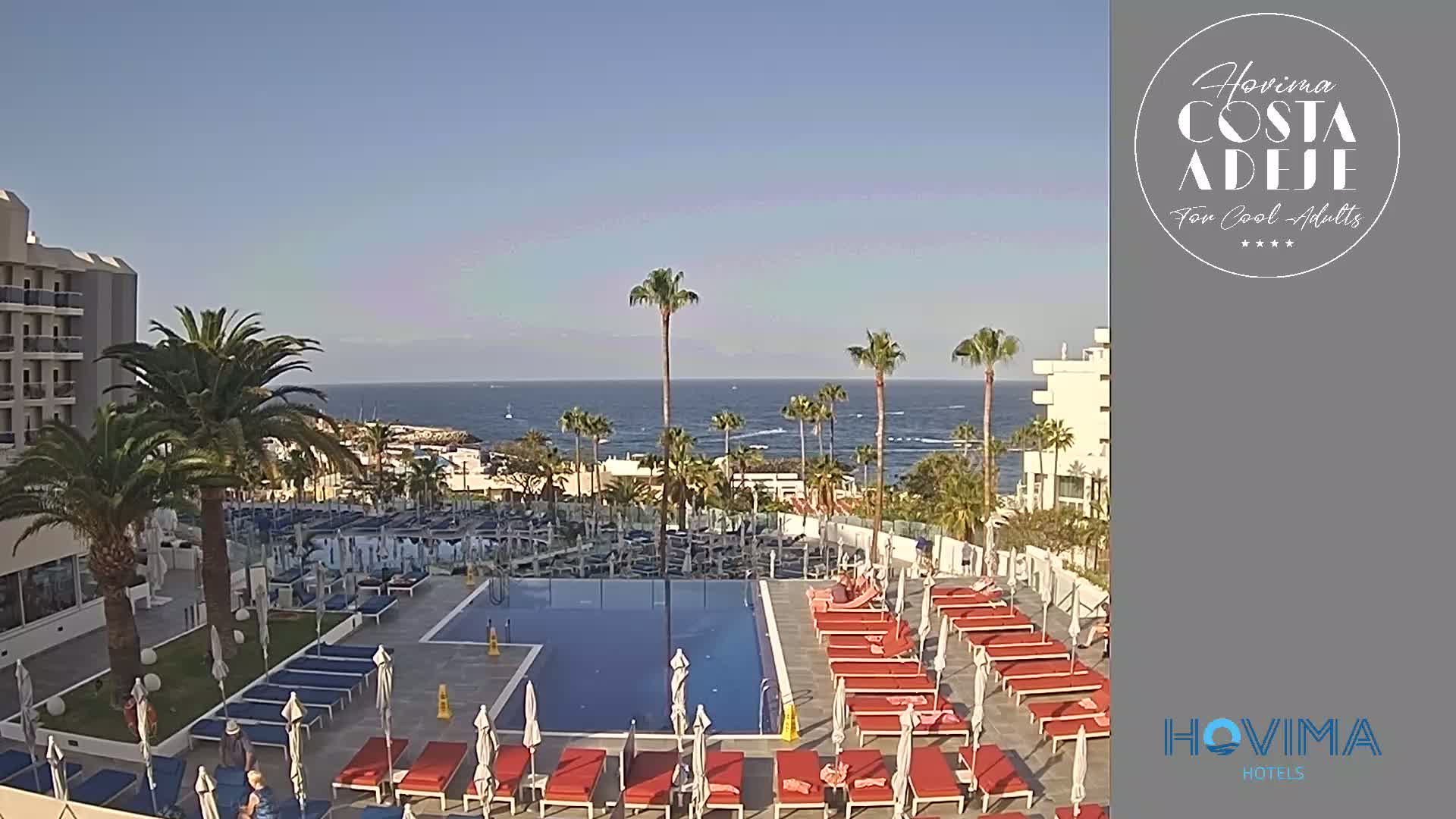 A hotel pool area with red lounge chairs and umbrellas surrounds a rectangular pool, overlooking a calm ocean under a clear blue sky.