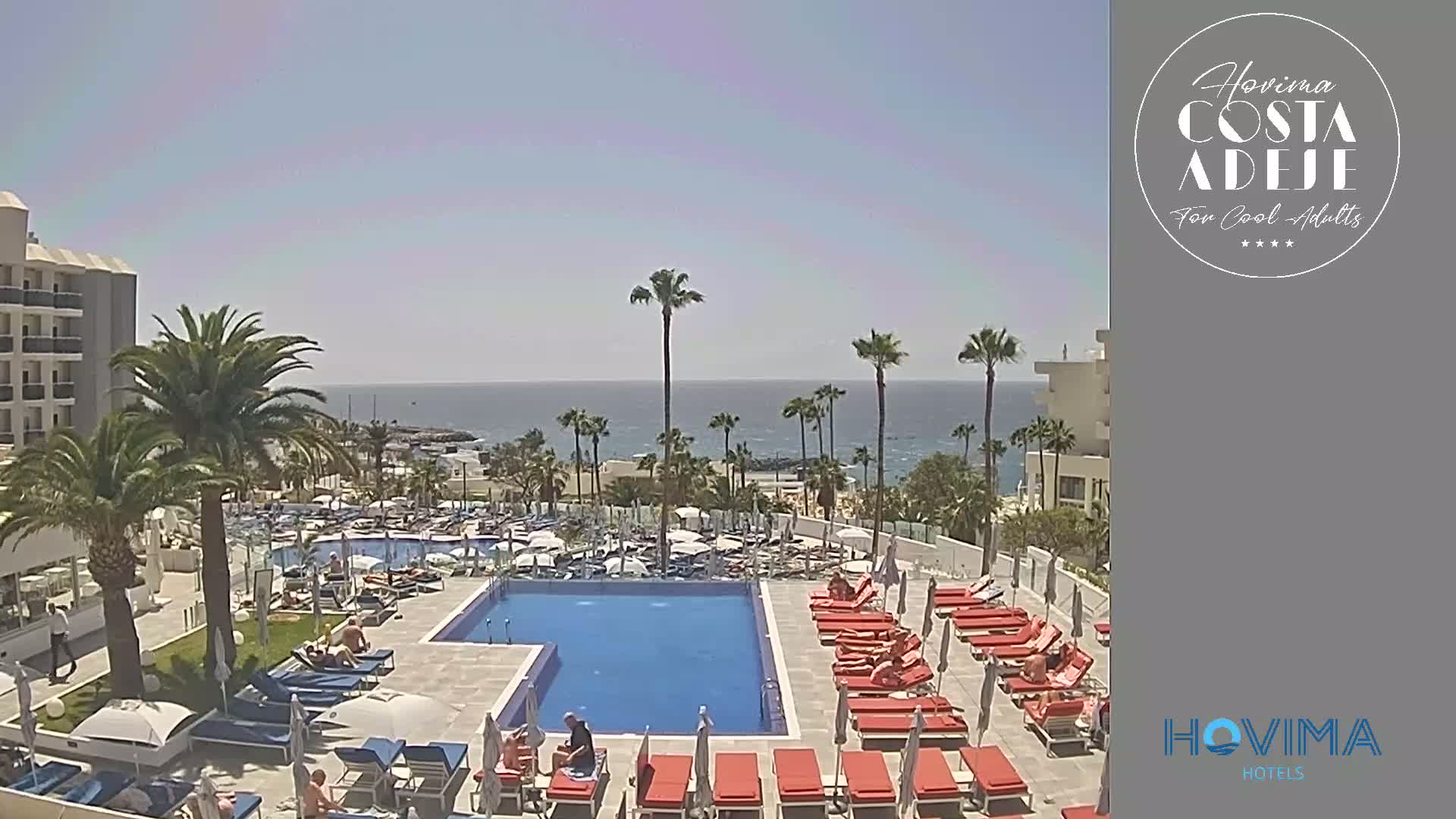 An outdoor pool area with multiple lounge chairs and umbrellas sits next to a hotel building, overlooking a calm ocean on a sunny day.