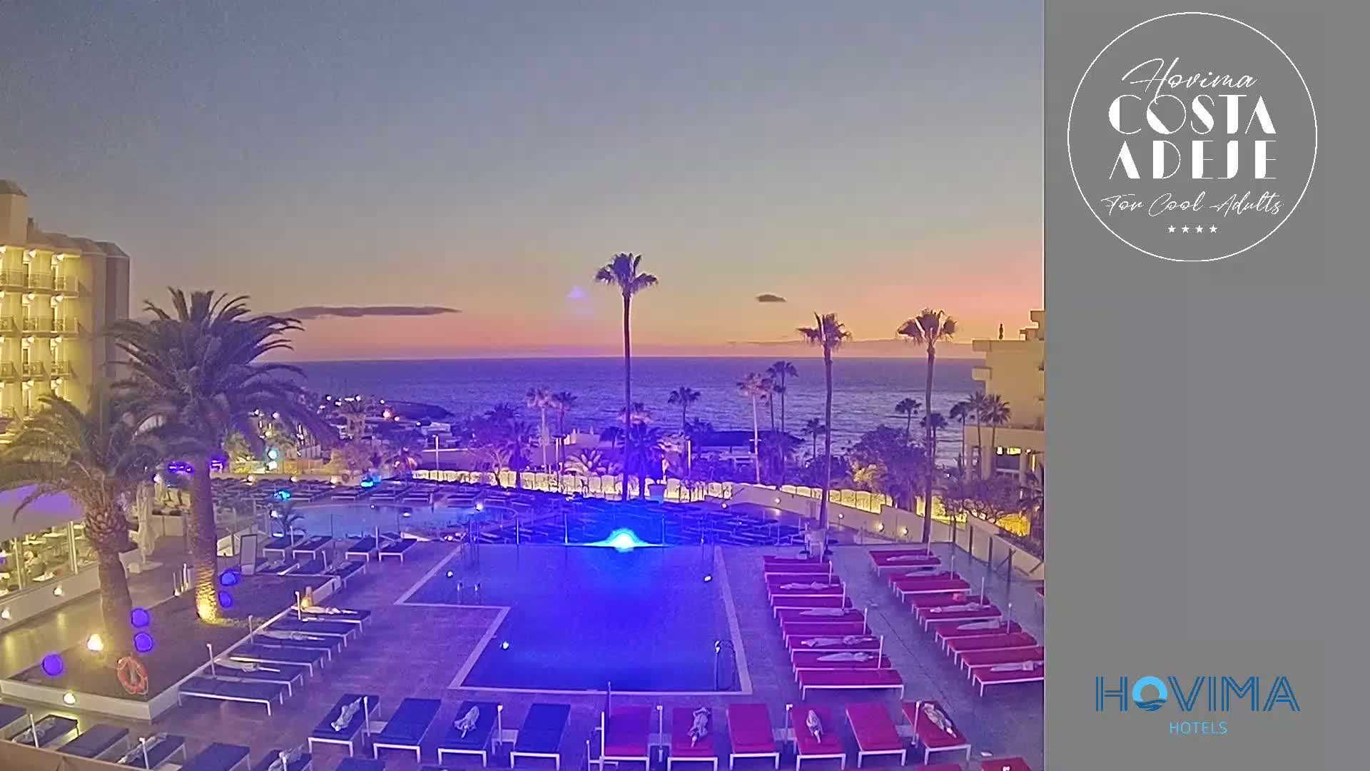 An illuminated pool area with numerous lounge chairs sits in front of a hotel overlooking a calm ocean at twilight.