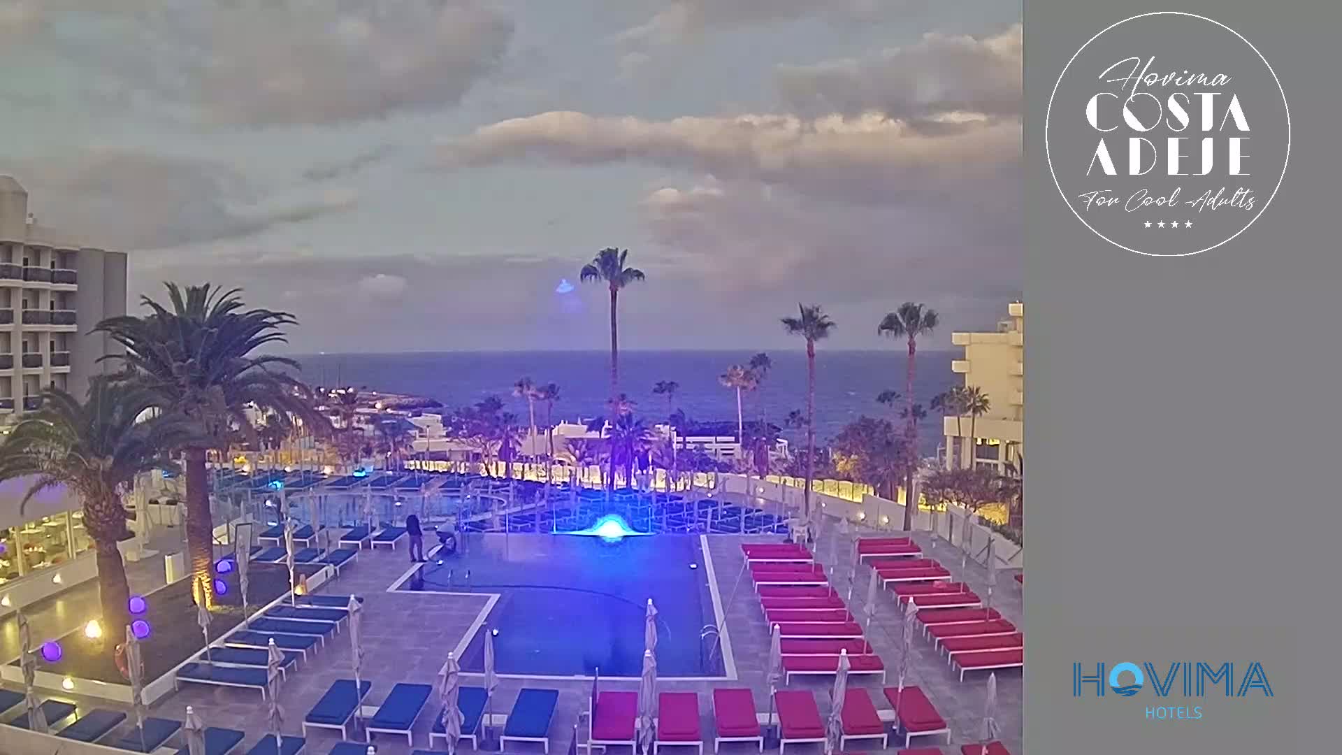 An outdoor pool area with lounge chairs and palm trees is illuminated at night under a cloudy sky, overlooking a calm ocean.