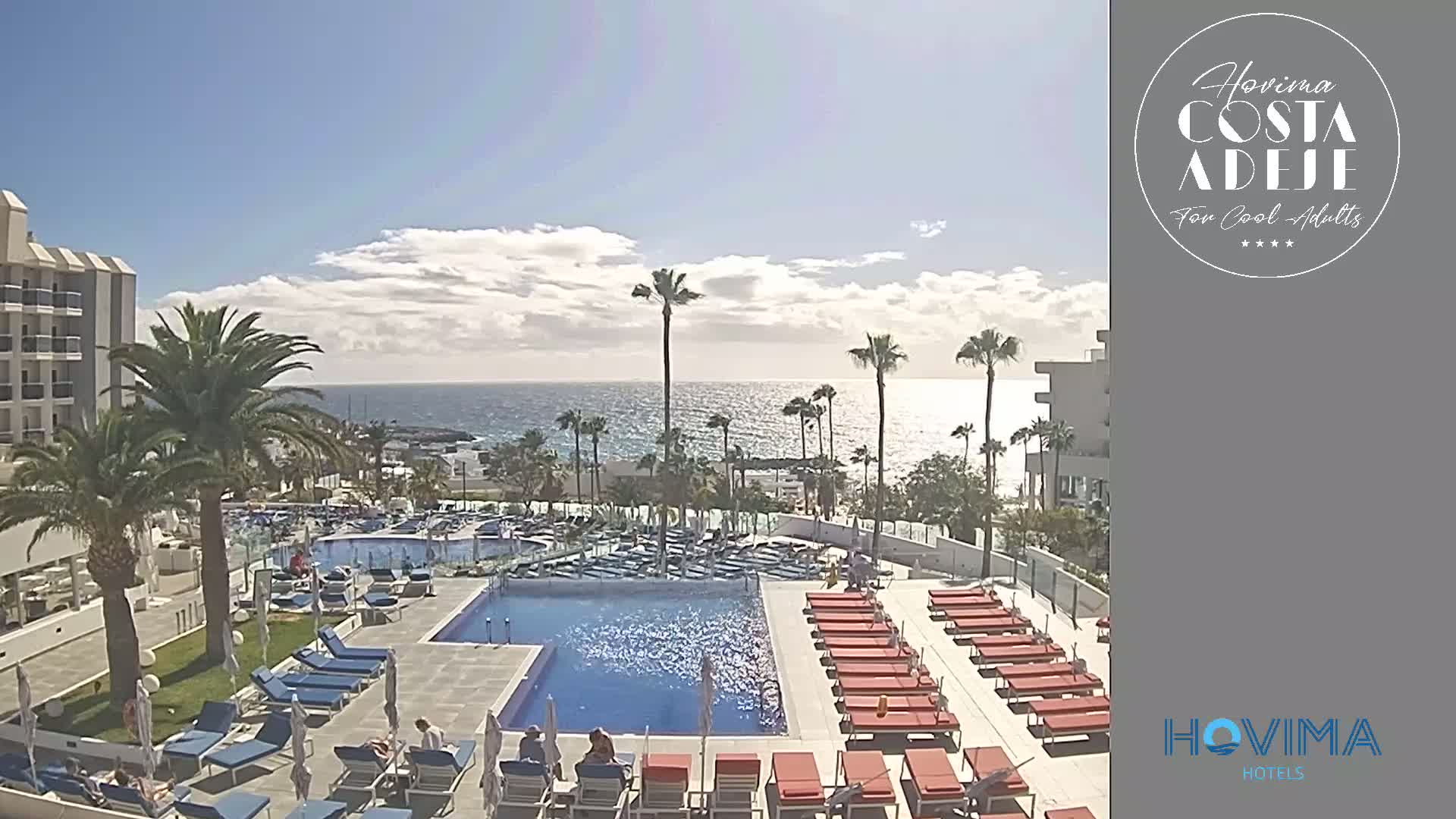 A resort pool area with lounge chairs surrounding a large pool, palm trees, and a view of a calm ocean under a partly cloudy sky.