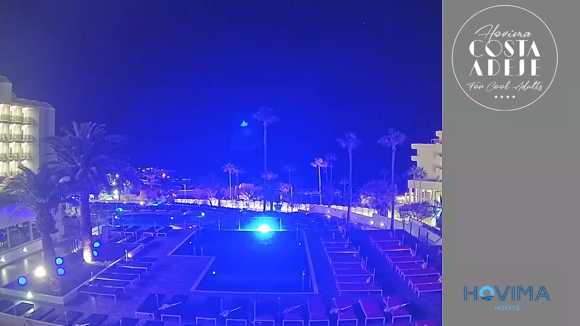 A nighttime view of a resort pool area with palm trees and a hotel building under a clear, dark blue sky.