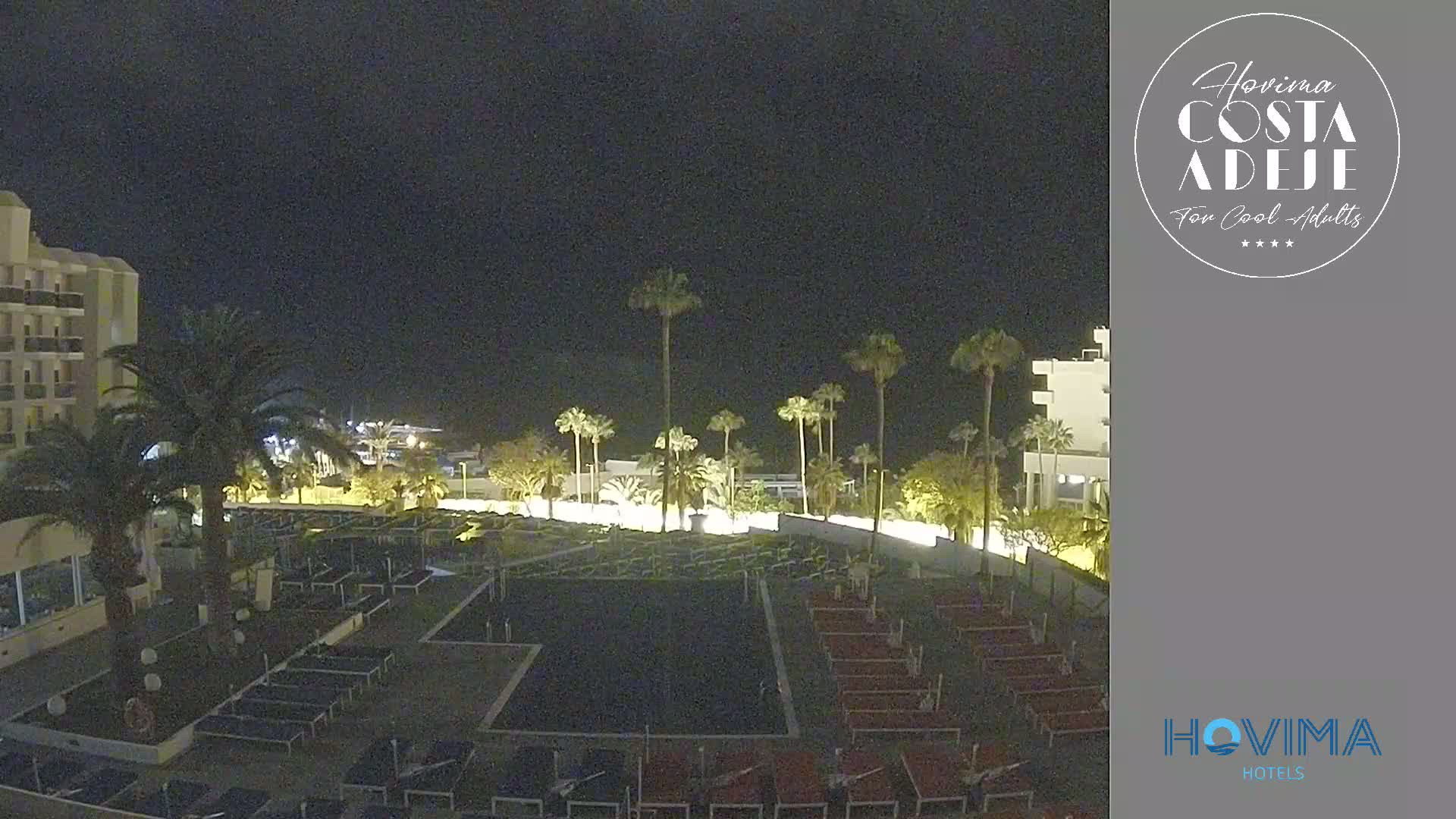 A nighttime view of a resort area with palm trees, buildings, and numerous lounge chairs arranged around a dark-colored court.
