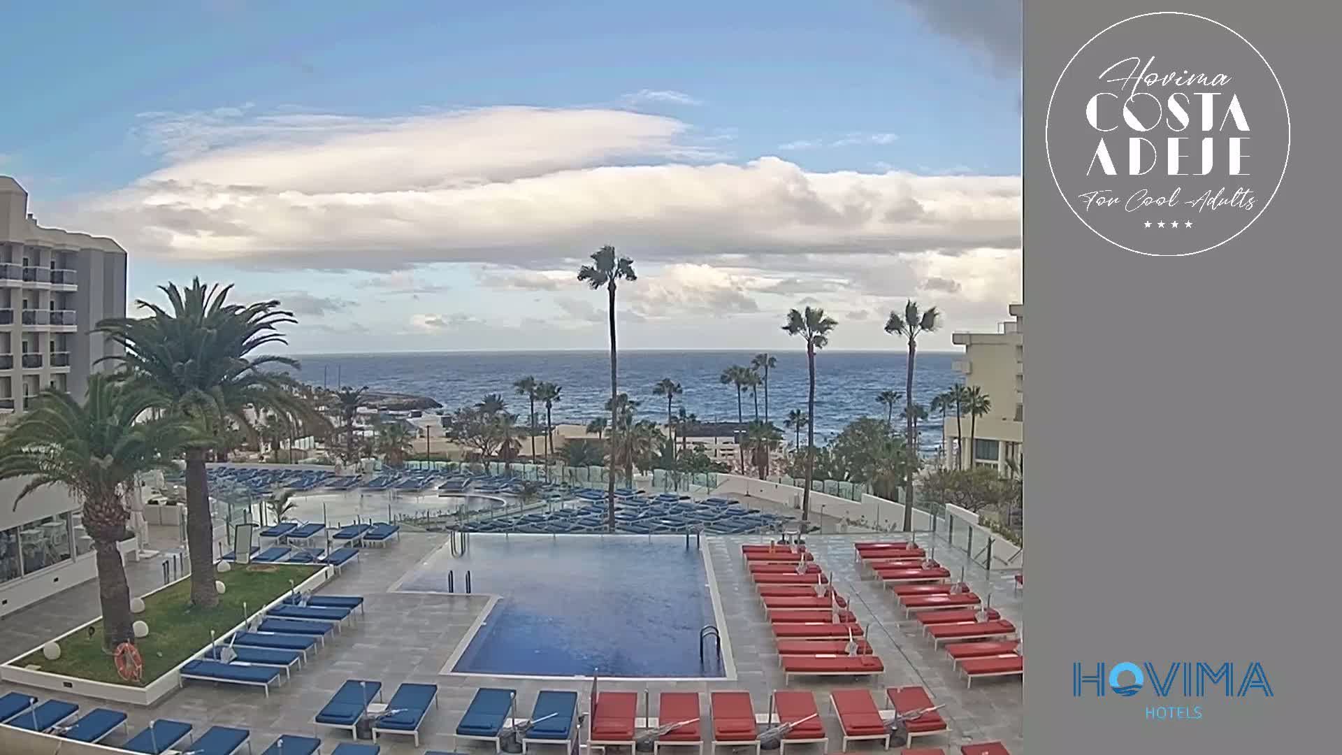 A resort pool area with numerous lounge chairs surrounds a rectangular pool, overlooking a calm ocean under a partly cloudy sky.