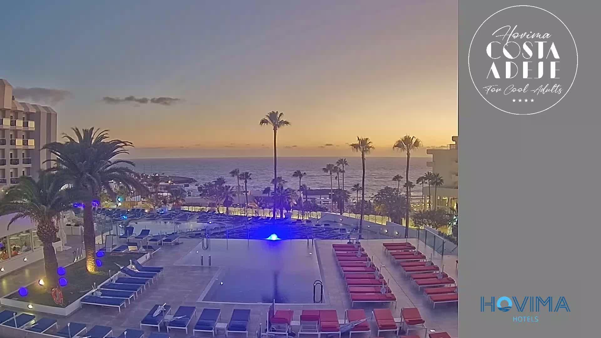 A twilight view of an outdoor swimming pool area at a resort, with palm trees, lounge chairs, and an ocean in the background.