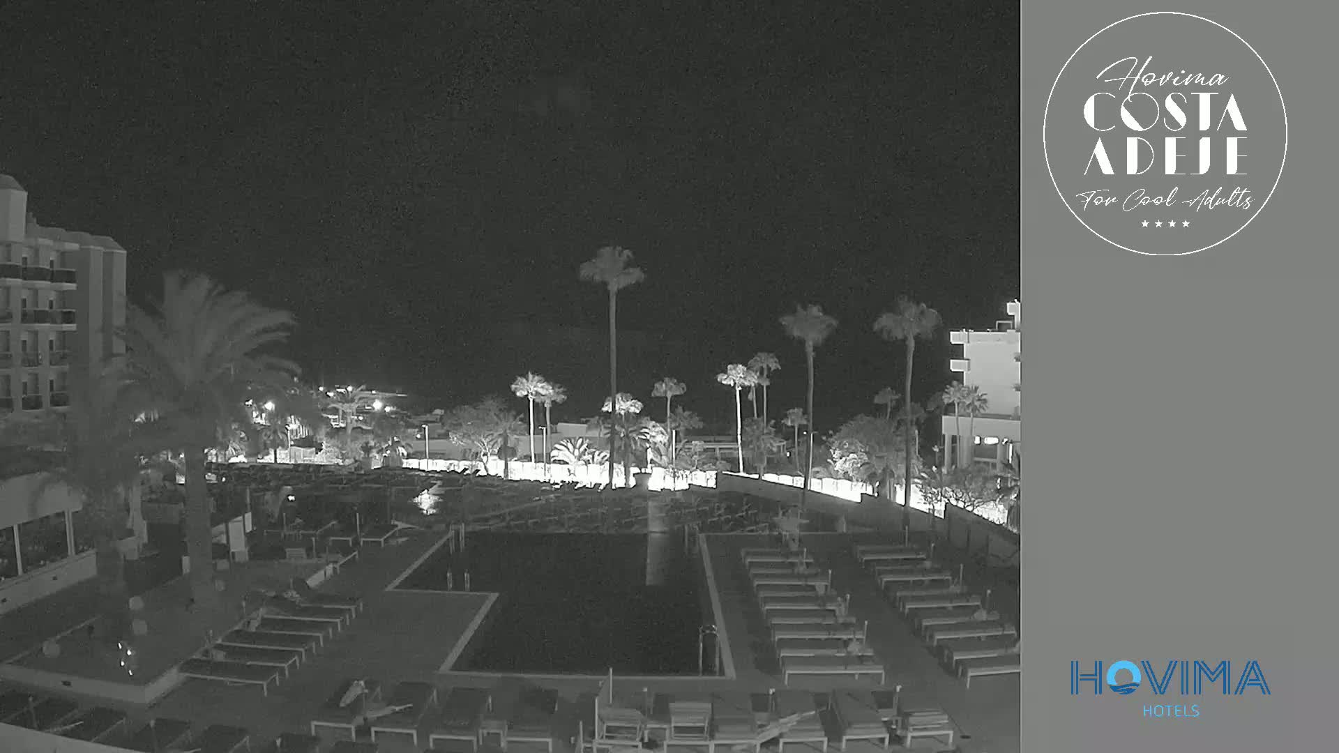 A nighttime view of a resort pool area with surrounding palm trees and buildings under a clear, dark sky.