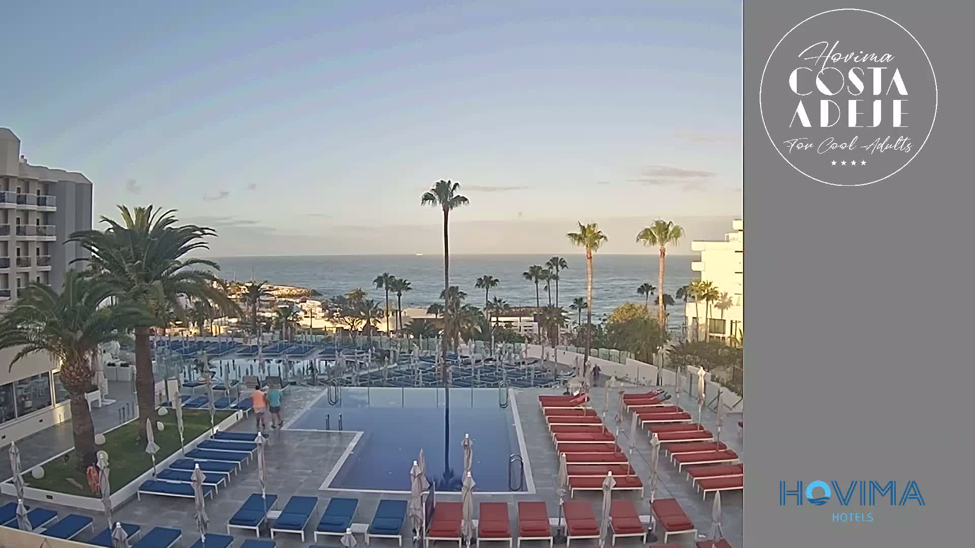 A resort pool area with lounge chairs and umbrellas overlooks a calm ocean under a clear sky.