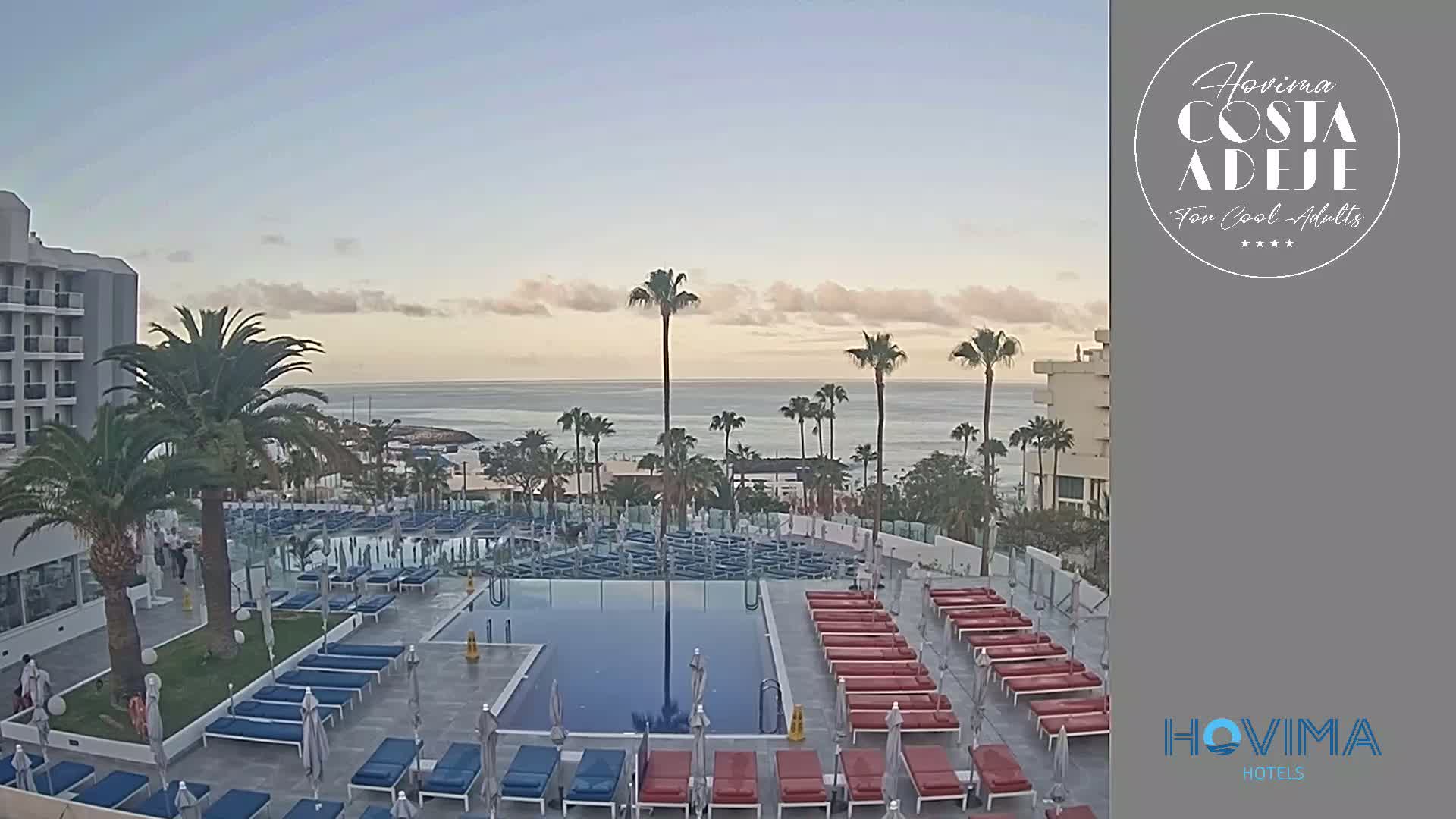 A hotel pool area with numerous lounge chairs surrounding a rectangular pool overlooks a calm ocean under a mostly clear sky.