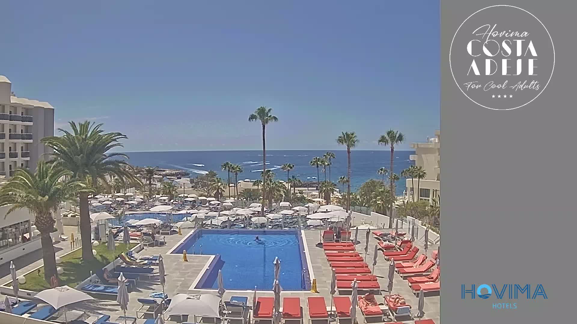 A resort pool area with red lounge chairs surrounds a blue pool, overlooking a calm ocean under a clear blue sky.