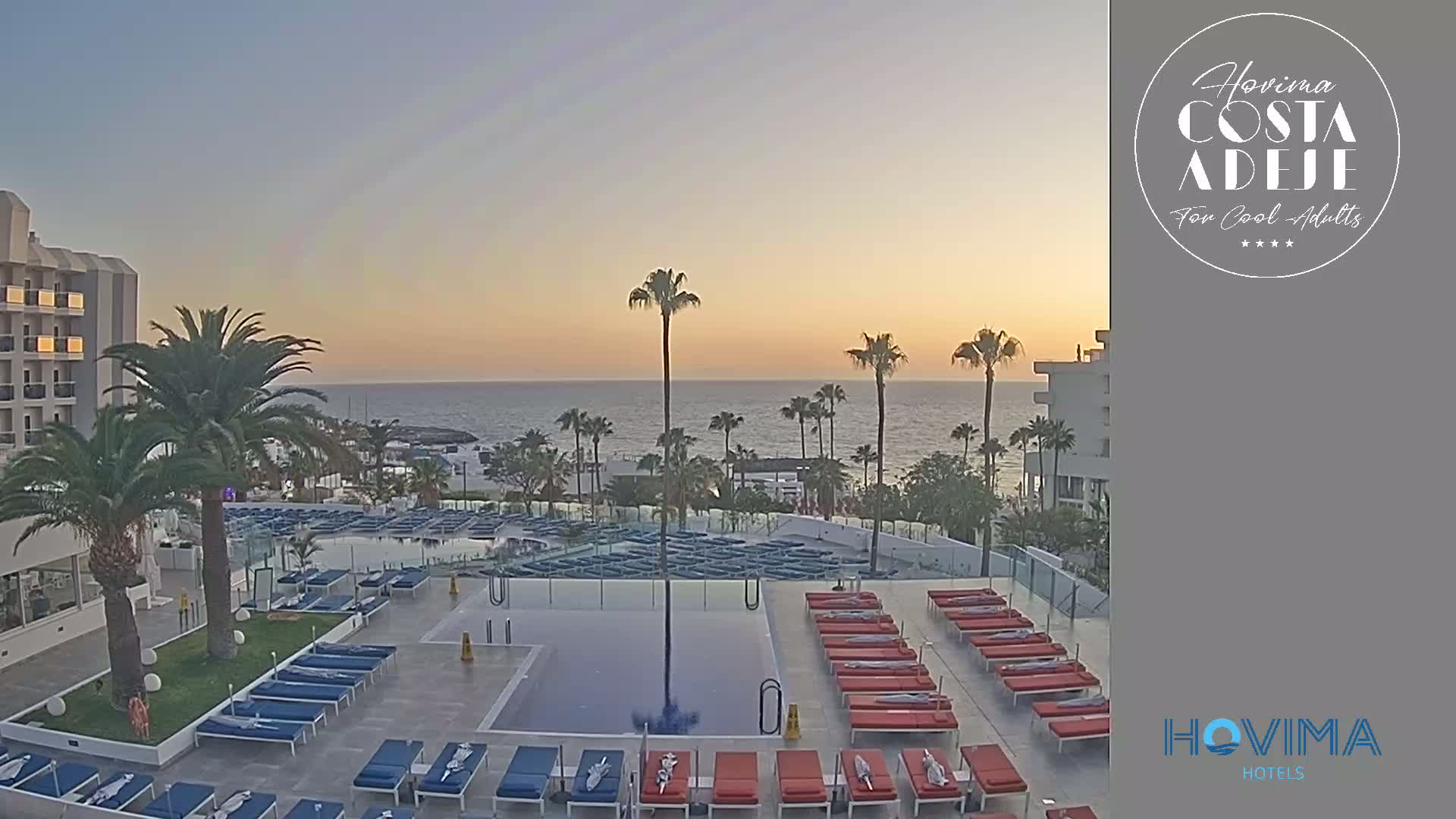 An outdoor pool area with numerous lounge chairs surrounding a rectangular pool, overlooks a calm ocean during a sunset.