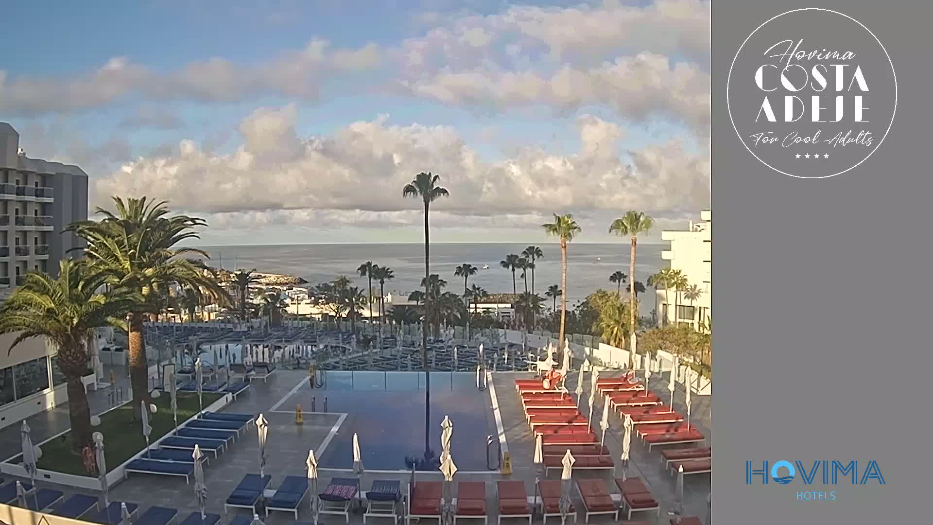 A resort pool area with lounge chairs and umbrellas sits below a hotel building overlooking a calm ocean under a partly cloudy sky.