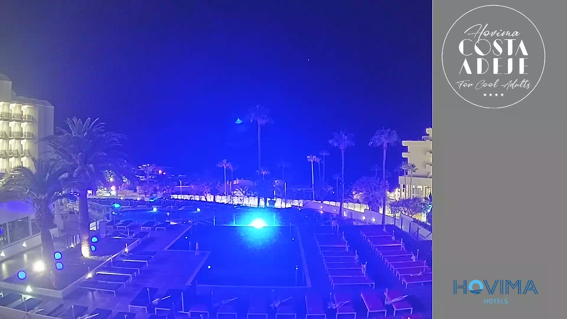 A nighttime view of a resort pool area illuminated in blue, surrounded by palm trees and a multi-story building under a clear, dark sky.