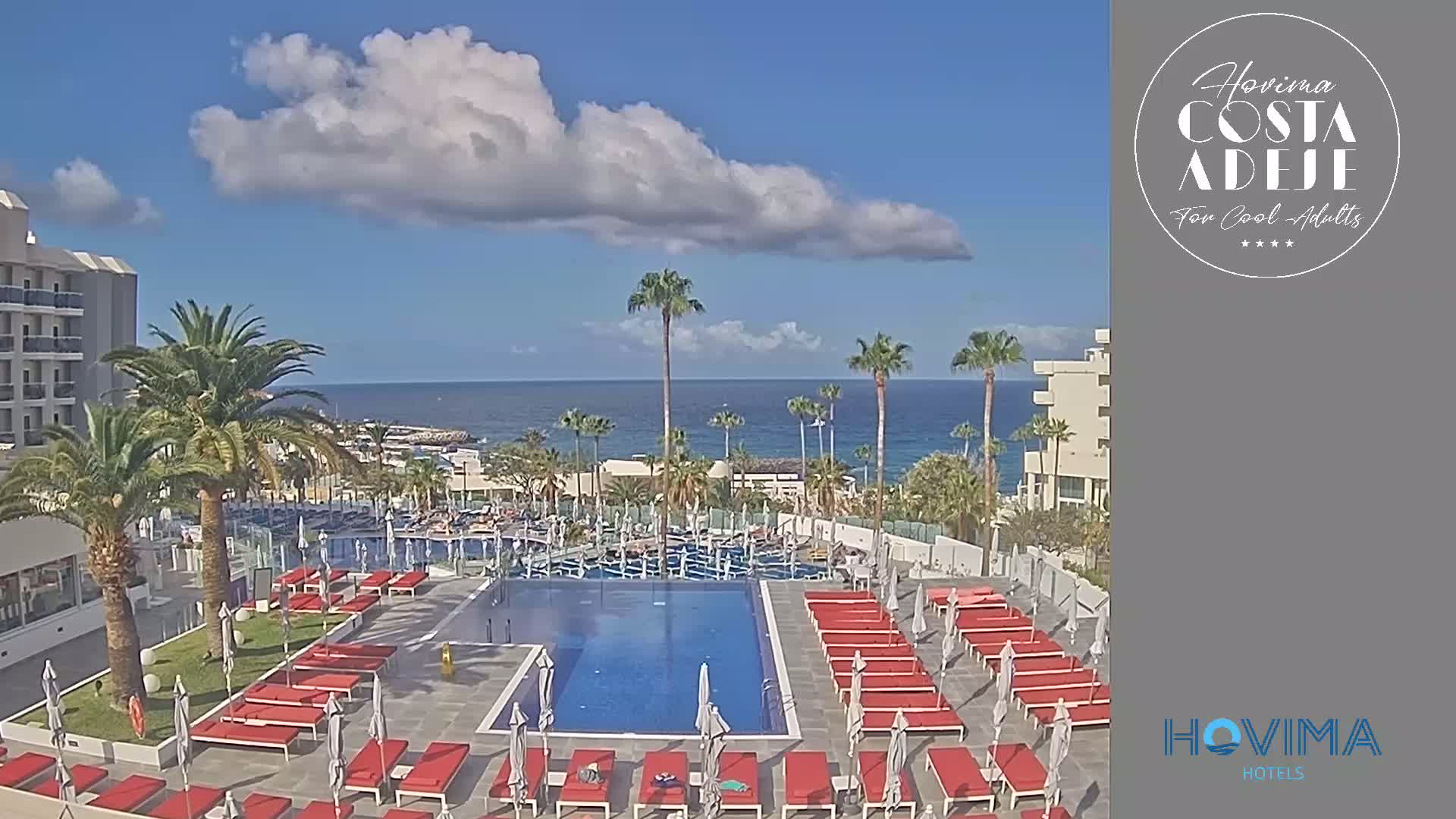 A resort pool area with red lounge chairs and umbrellas sits below a modern hotel building overlooking a calm ocean under a partly cloudy sky.