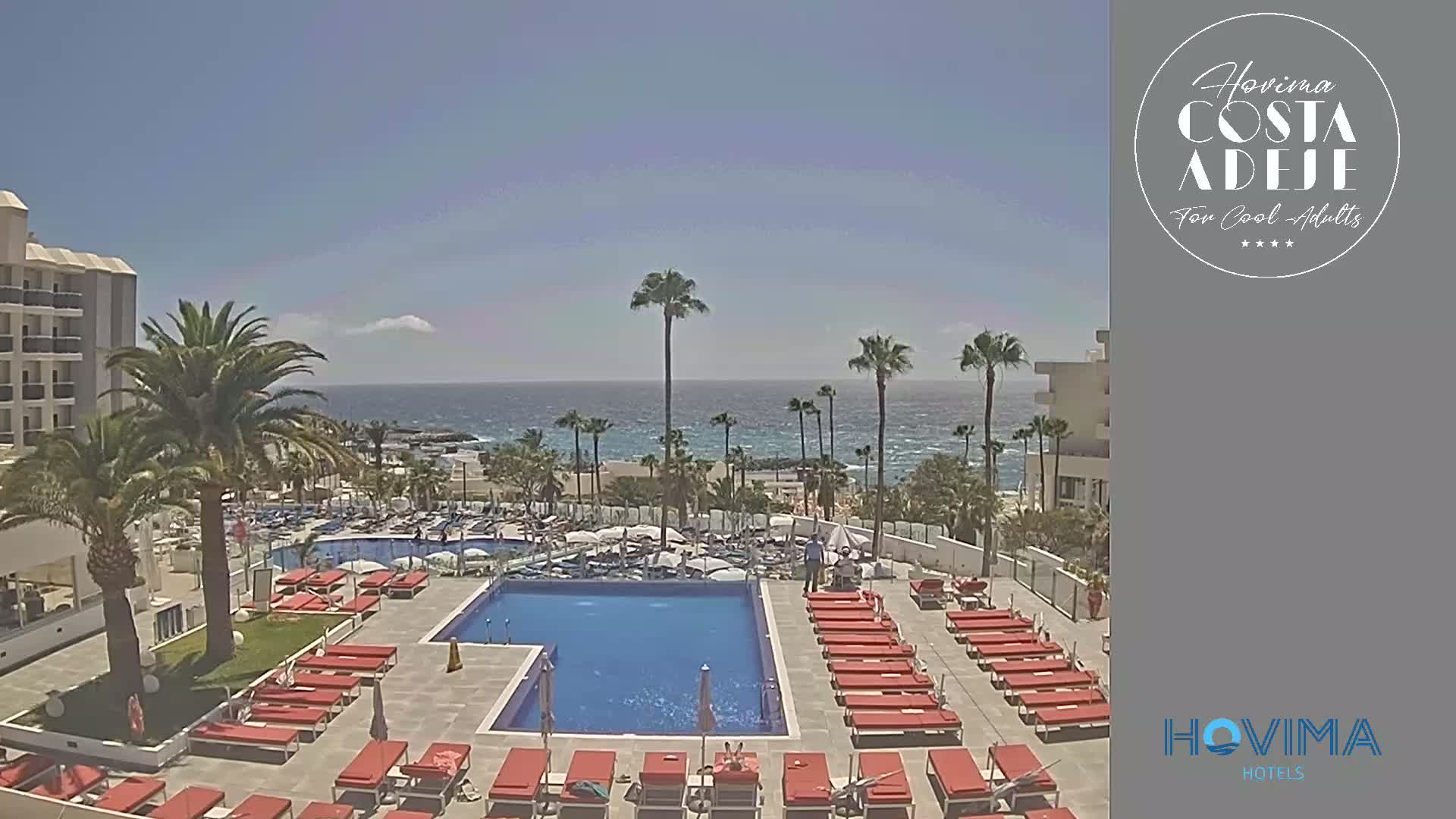 A resort pool area with red lounge chairs surrounds a large rectangular pool, overlooking a beach and ocean under a clear, sunny sky.