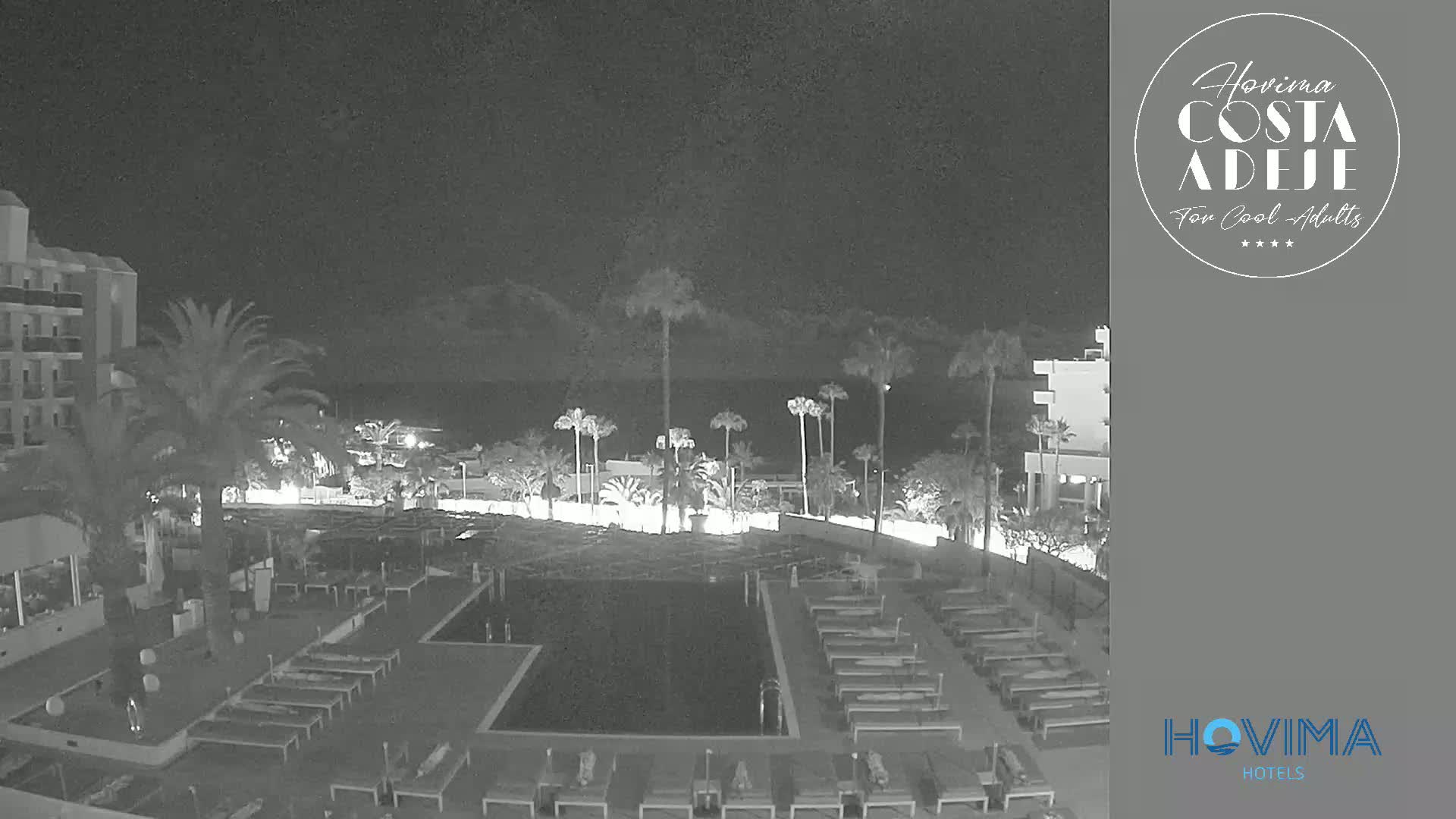 A nighttime view of a resort pool area with palm trees, lounge chairs, and a building, overlooking a dark body of water under a clear sky.
