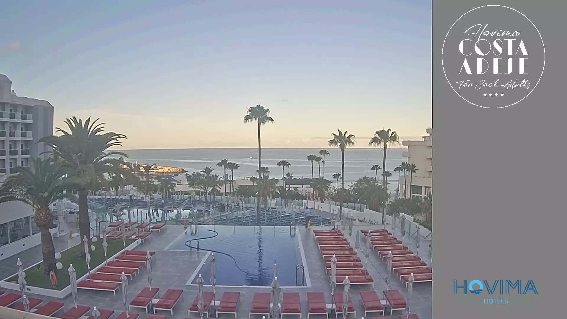 A resort pool area with red lounge chairs and umbrellas overlooks a calm ocean under a clear sky.