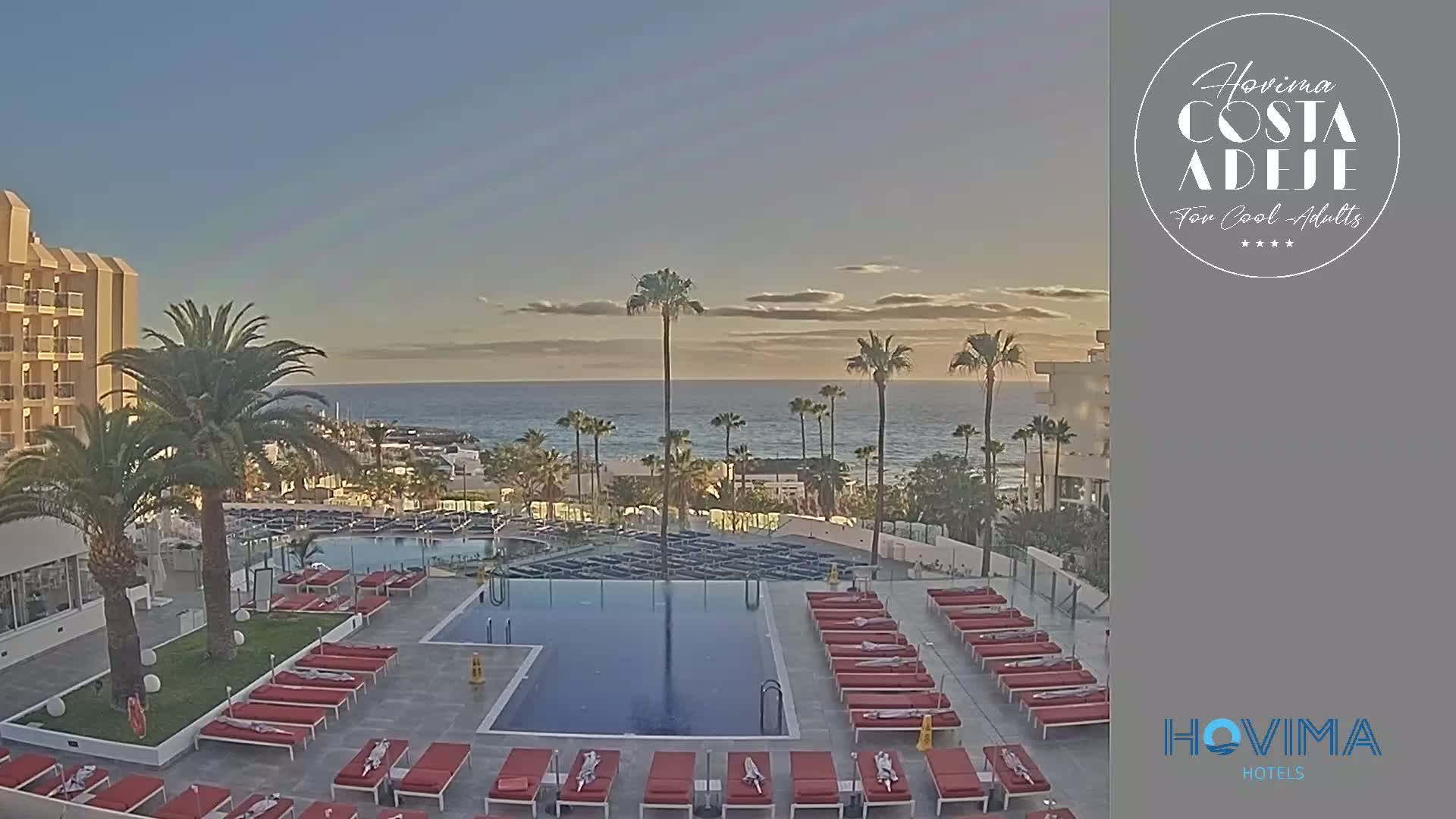 A resort pool area with red lounge chairs surrounds a rectangular pool, overlooking a calm ocean at sunset under a clear sky.
