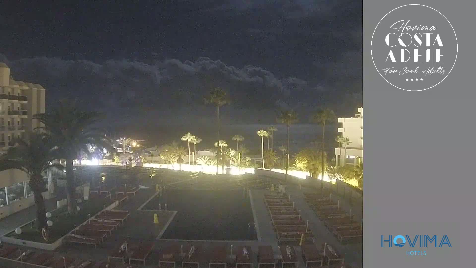 A nighttime view of a resort area with palm trees, a pool, and rows of lounge chairs under a cloudy sky.