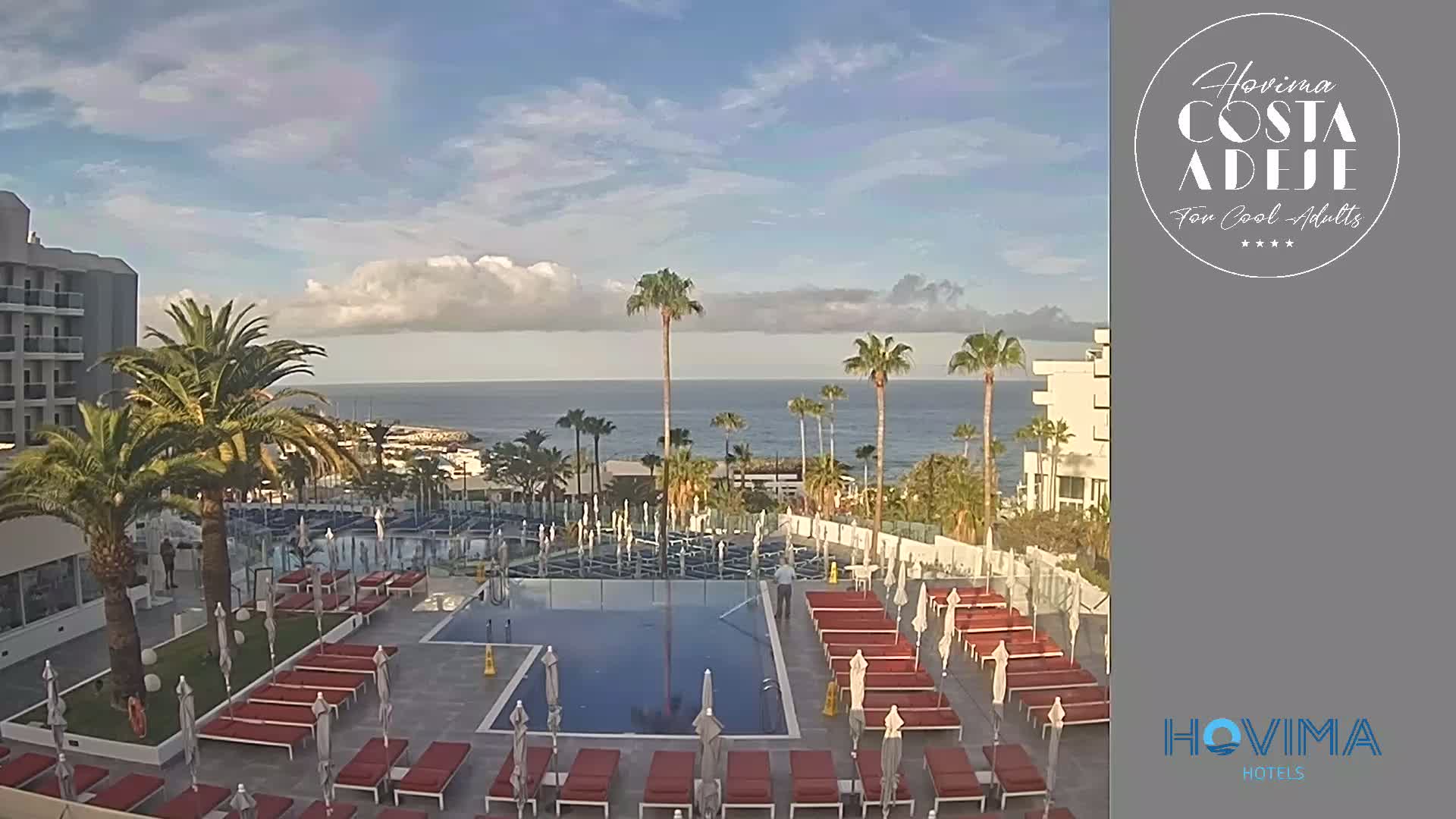 A resort pool area with red lounge chairs and umbrellas overlooks a calm ocean under a partly cloudy sky.
