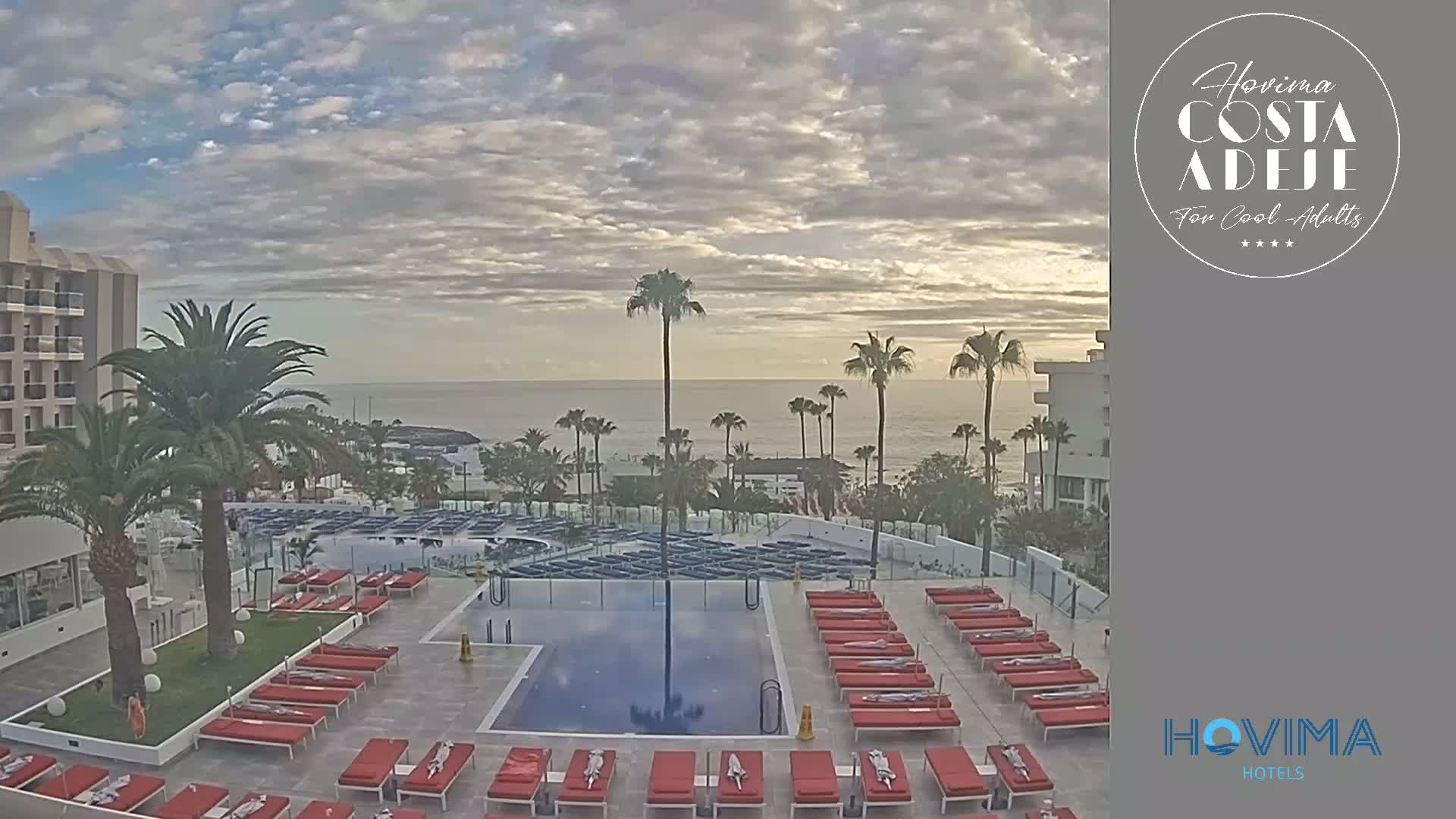 An outdoor pool area with red lounge chairs sits adjacent to a hotel overlooking a calm ocean under a cloudy sky.