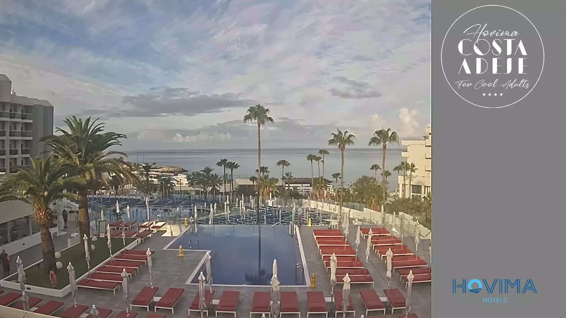 An outdoor swimming pool with red lounge chairs and umbrellas is situated next to a hotel, overlooking a calm ocean under a partly cloudy sky.