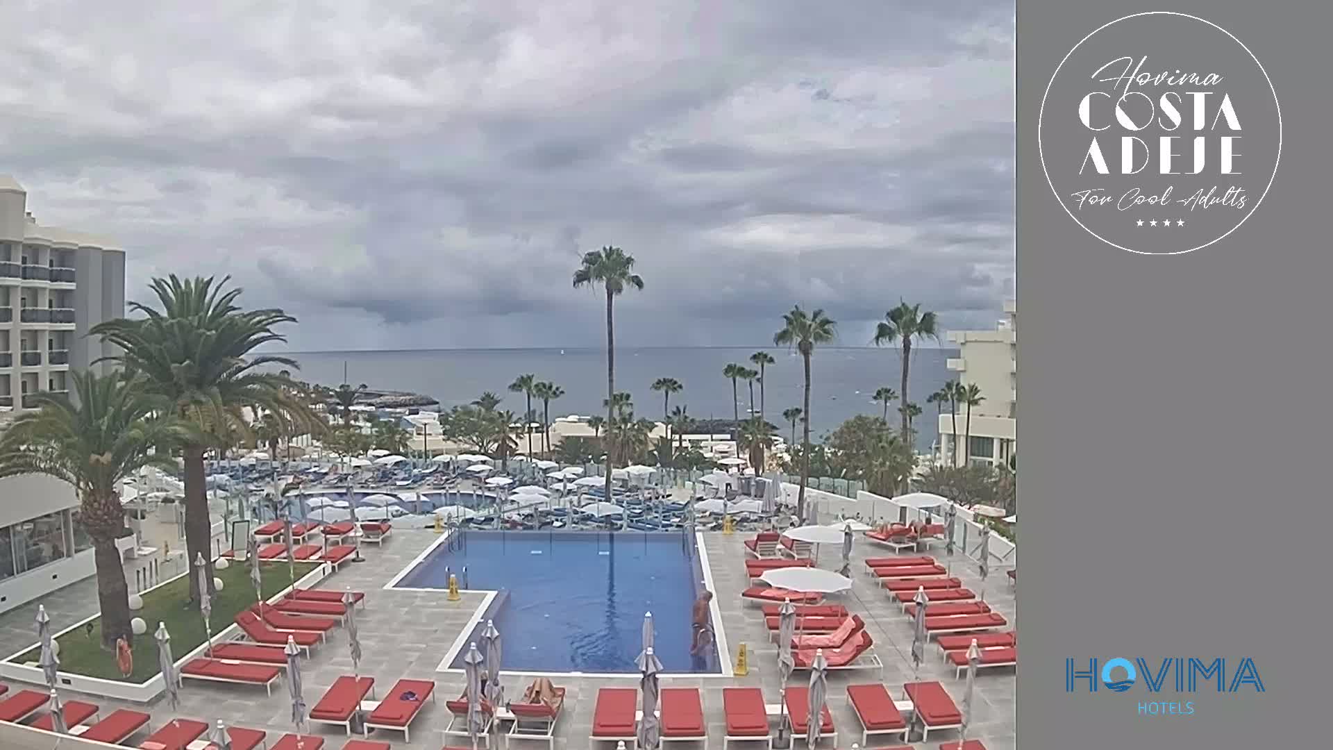 An outdoor pool area with red lounge chairs and palm trees overlooks a calm ocean under a cloudy sky.