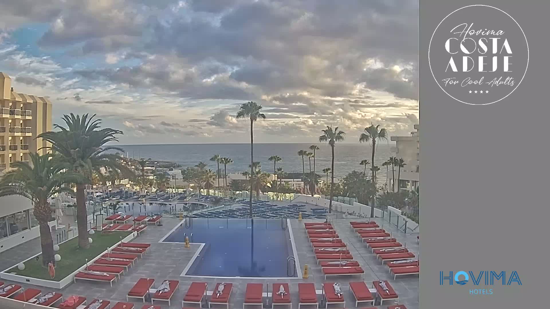 A hotel pool area with numerous red lounge chairs surrounds a rectangular pool, overlooking a calm ocean under a partly cloudy sky.