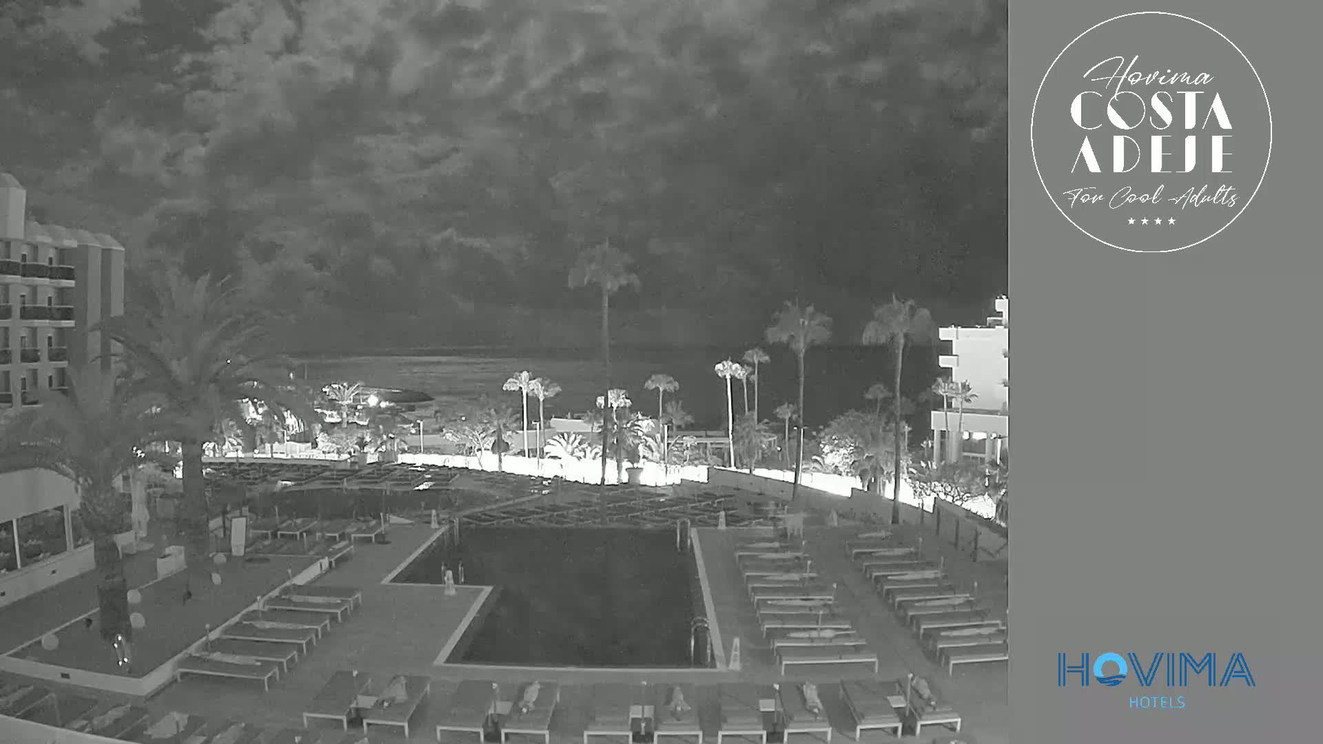 A nighttime view of a resort pool area with palm trees and a building under a cloudy sky.