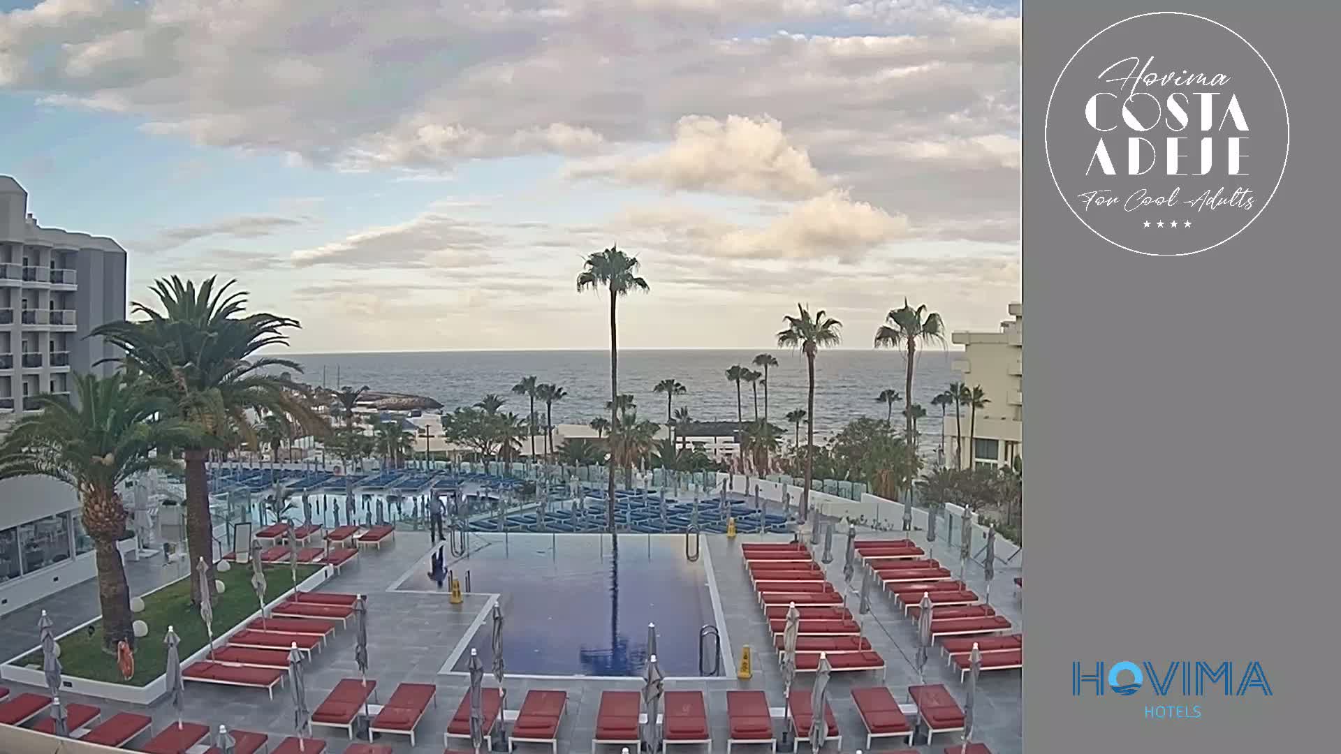 An outdoor pool area with many red lounge chairs surrounds a rectangular pool; palm trees and a building are visible, and a calm ocean is in the background under a partly cloudy sky.