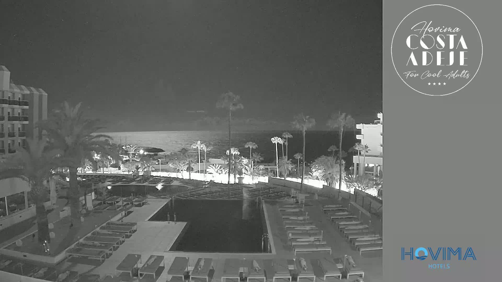 A nighttime, low-light view of a resort pool area with lounge chairs surrounding it, palm trees lining the perimeter, and a calm ocean in the background.