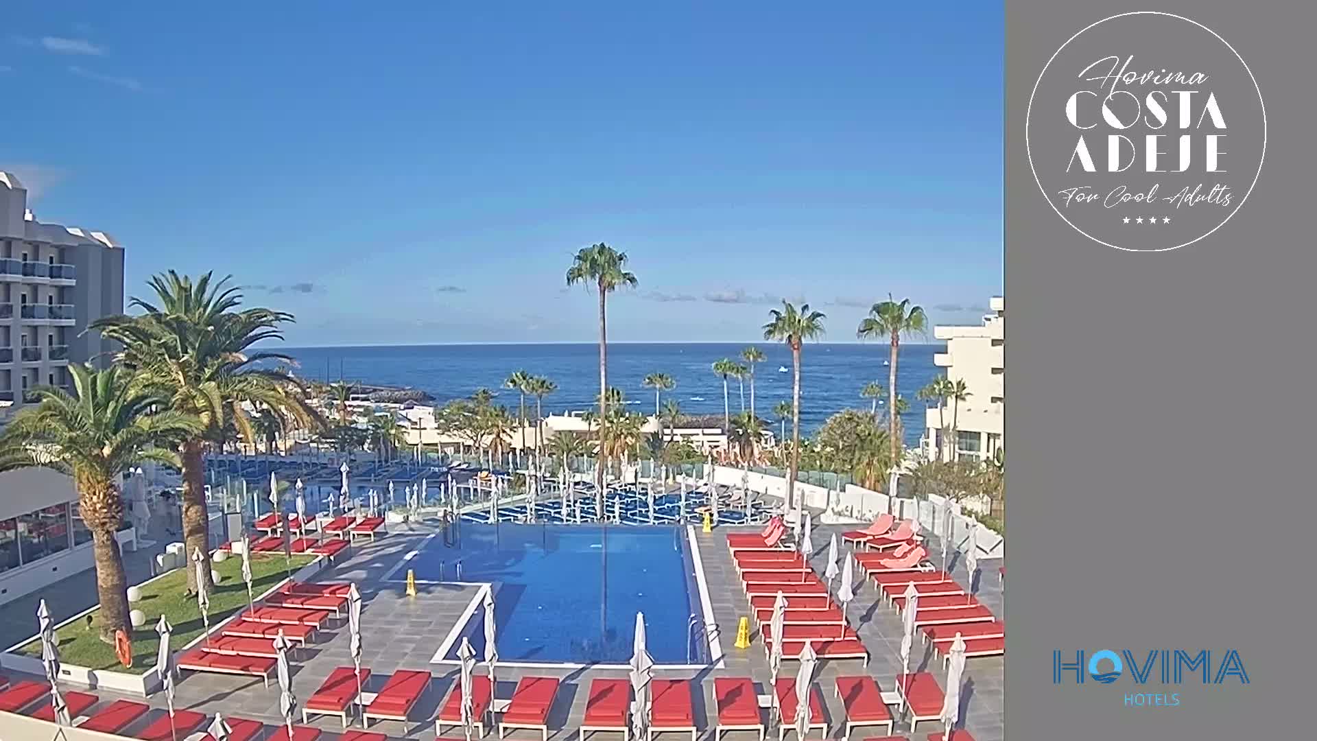 A resort pool area with red lounge chairs and umbrellas is situated next to a modern hotel building overlooking a calm ocean under a clear blue sky.