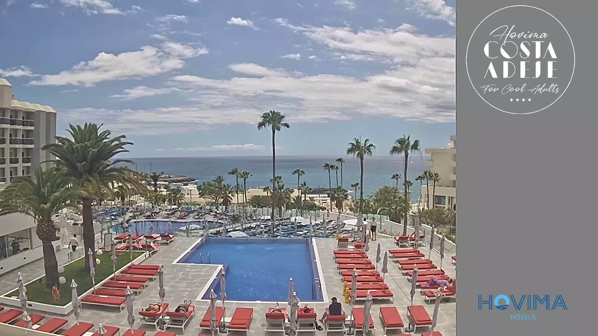 A resort pool area with red lounge chairs and umbrellas overlooks a calm ocean under a partly cloudy sky.