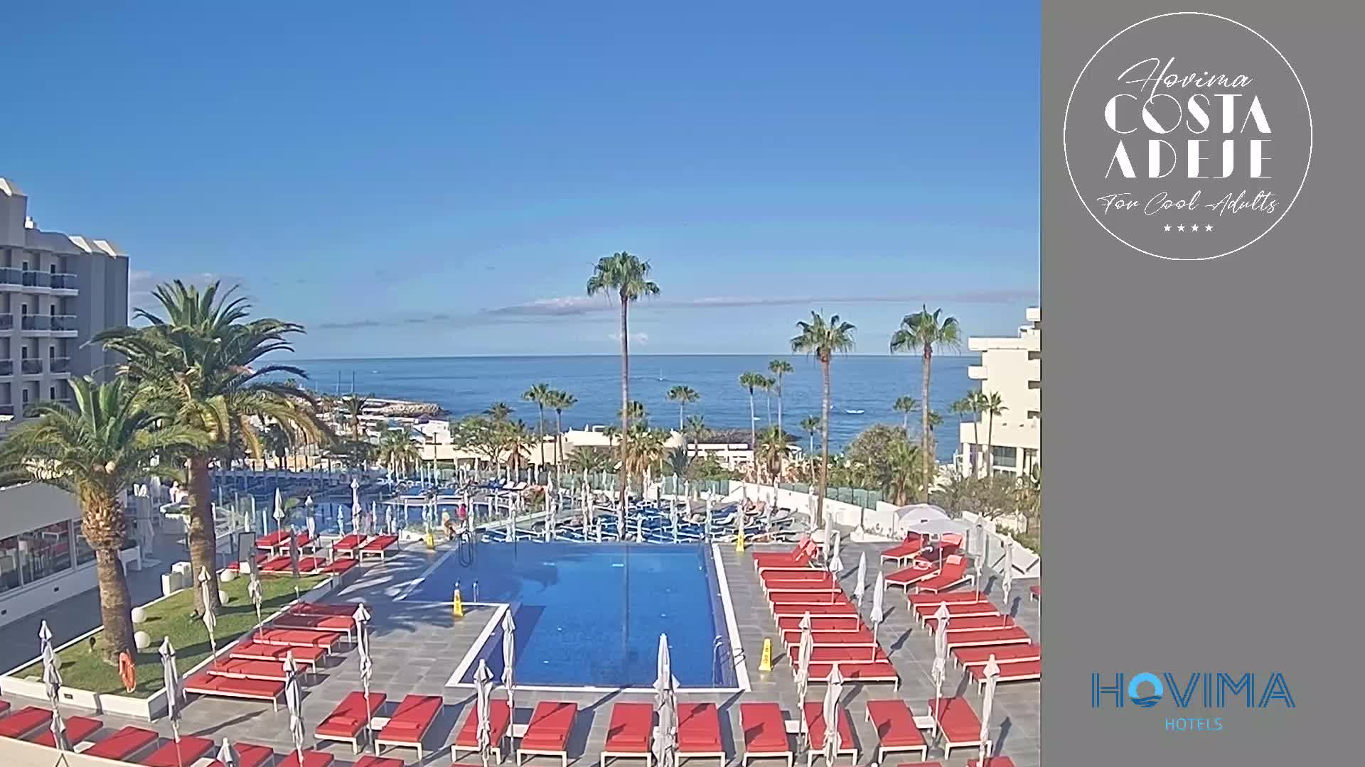 A resort pool area with red lounge chairs and umbrellas overlooks a calm ocean under a clear blue sky.