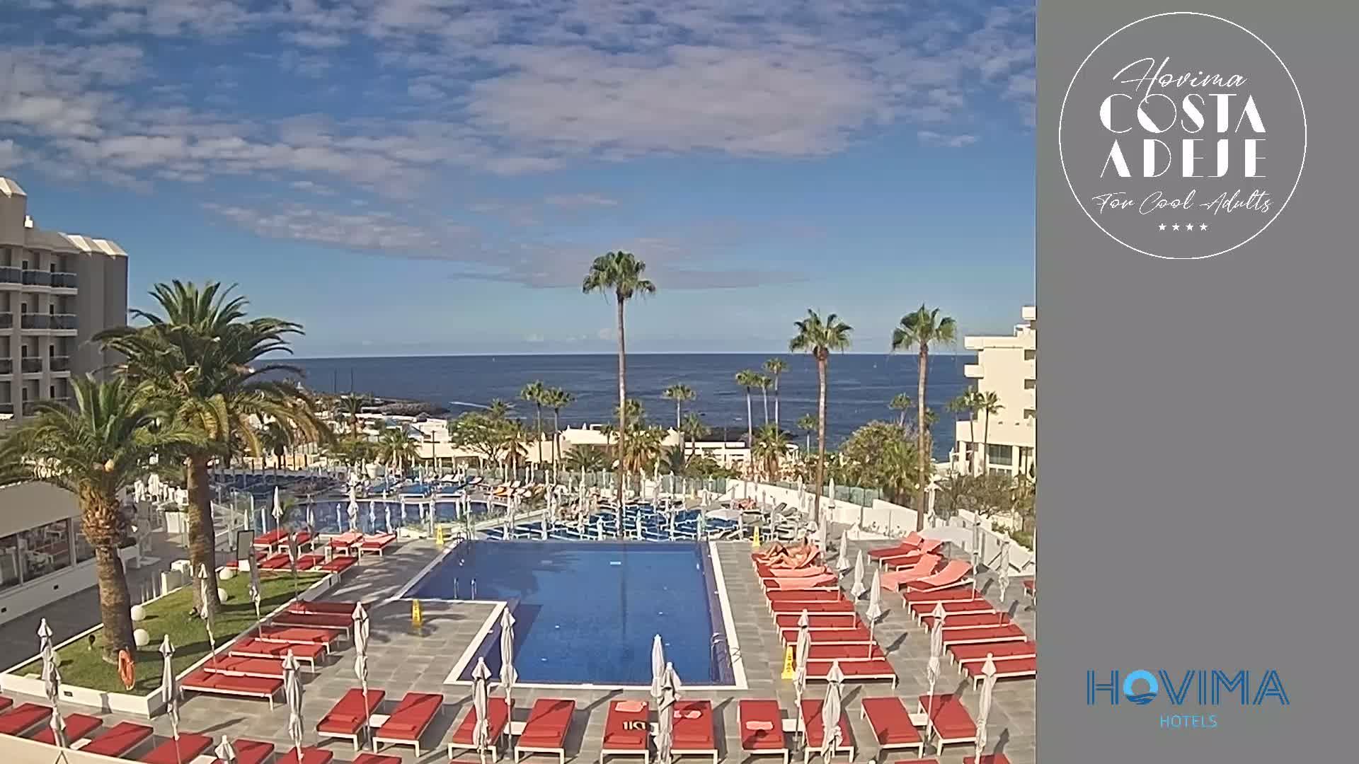 A resort pool area with red lounge chairs and umbrellas surrounds a large swimming pool, overlooking a calm ocean under a partly cloudy sky.