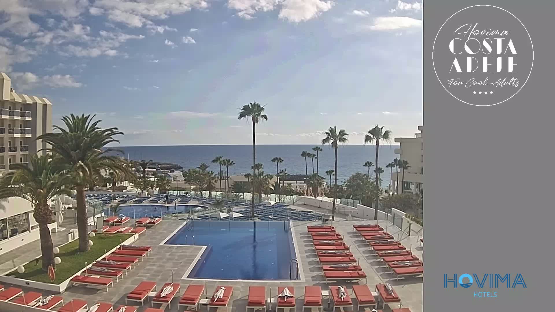 A hotel pool area with red lounge chairs surrounds a rectangular pool, overlooking a calm ocean under a partly cloudy sky, and palm trees line the perimeter.