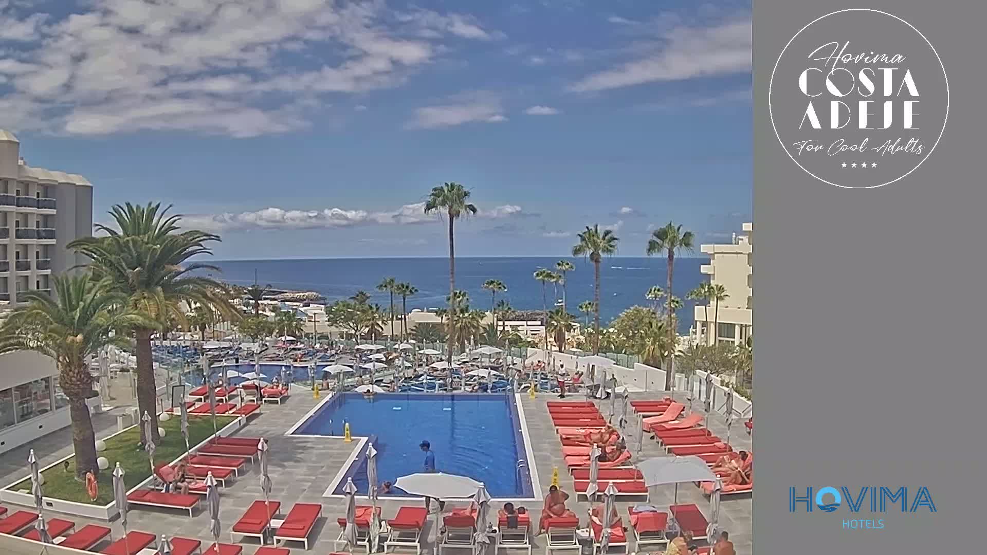 A resort pool area with red lounge chairs and umbrellas surrounds a large rectangular pool; palm trees and a building are visible, and a partly cloudy sky overlooks a calm ocean in the distance.