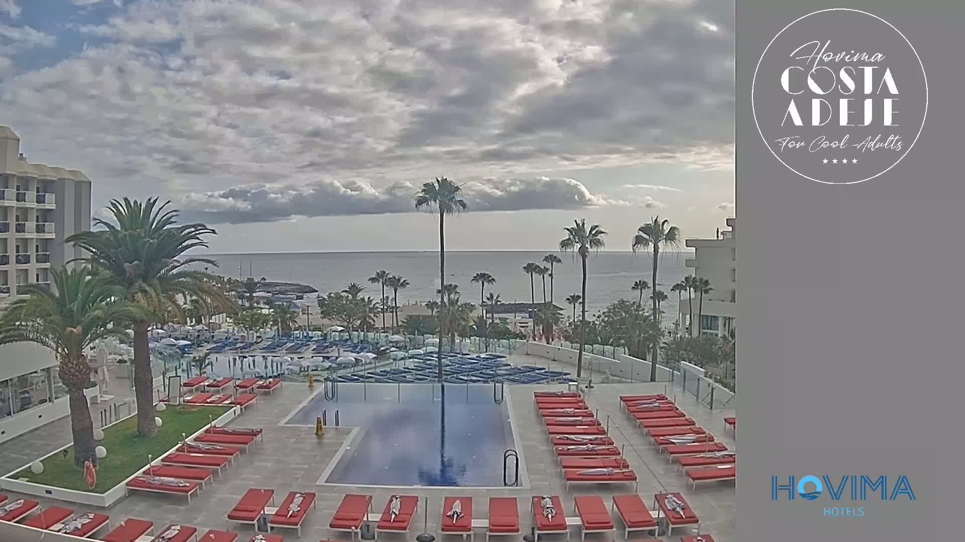 An outdoor pool with red lounge chairs is situated at a resort overlooking a calm ocean under a partly cloudy sky.