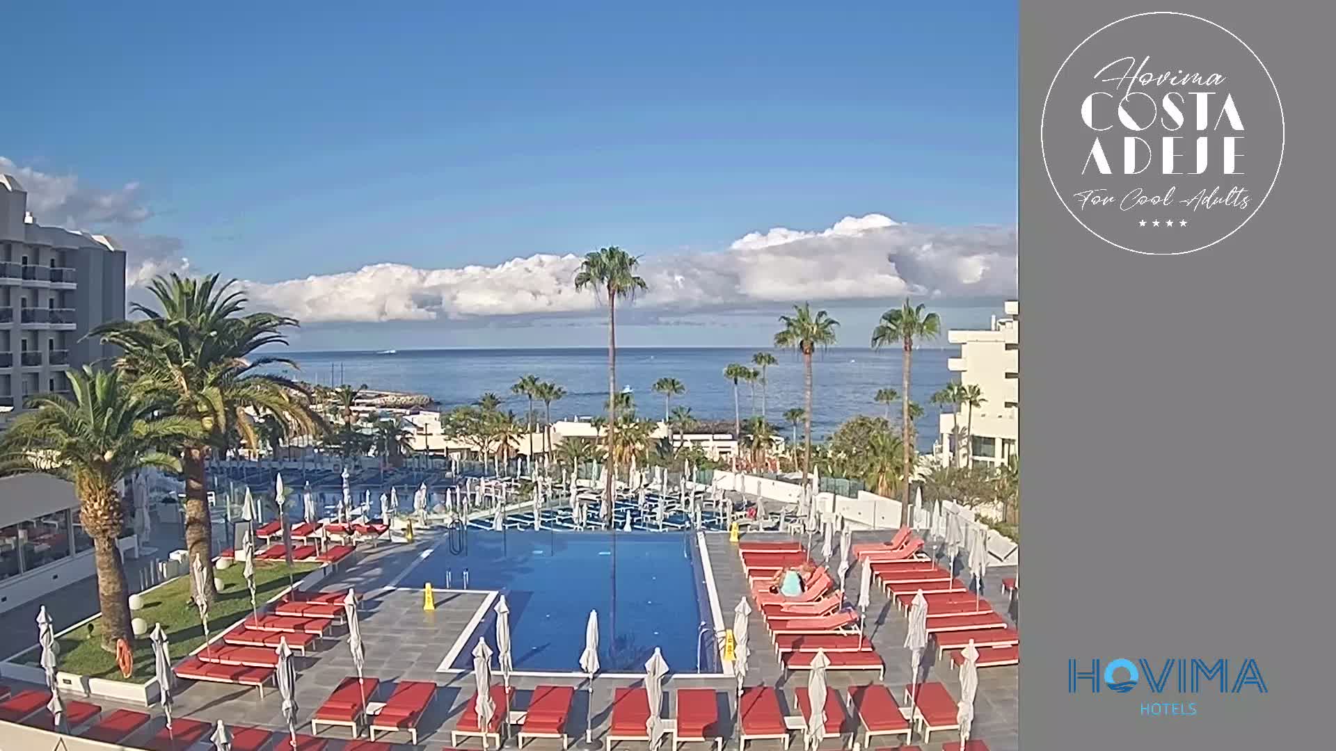 A resort pool area with red lounge chairs and umbrellas, palm trees, and a view of a calm ocean under a partly cloudy sky.