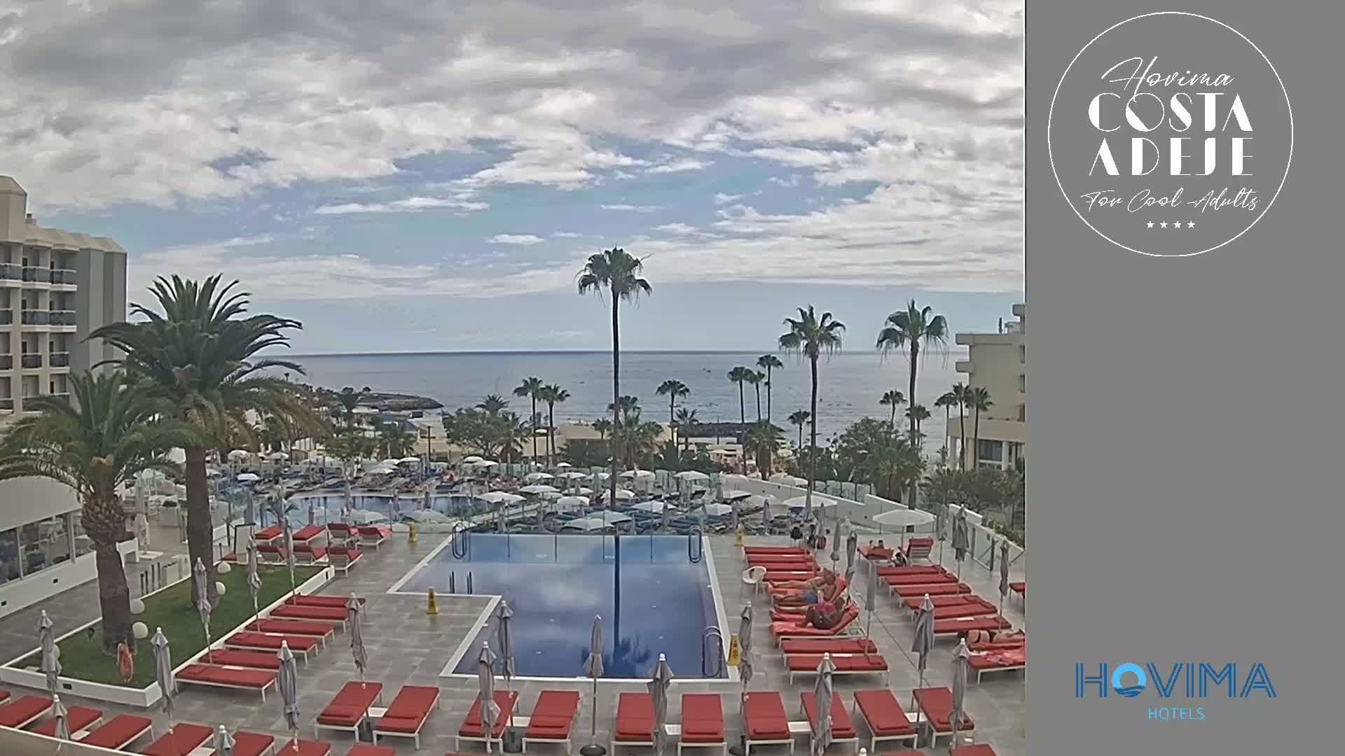 A resort pool area with red lounge chairs and umbrellas overlooks a calm ocean under a partly cloudy sky.