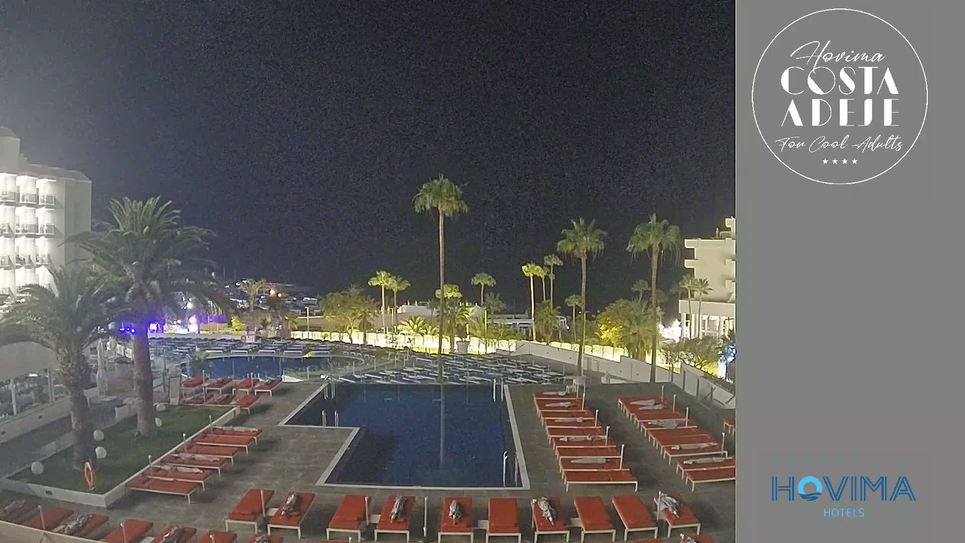 A nighttime view of a resort pool area with numerous empty lounge chairs surrounding a rectangular pool, palm trees in the background, and a multi-story hotel building to the left.