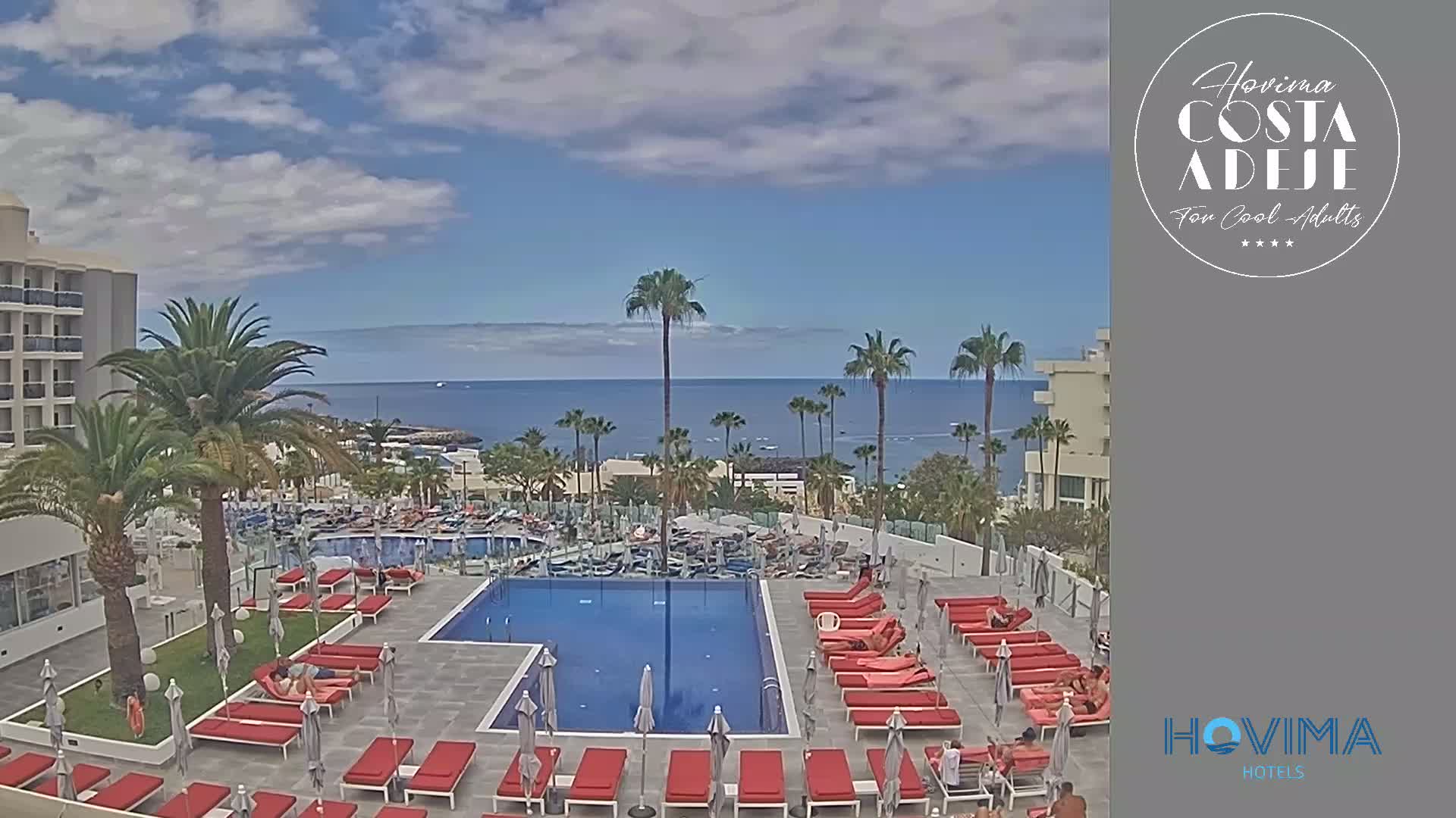An outdoor pool area with red lounge chairs and palm trees overlooks a calm ocean under a partly cloudy sky.