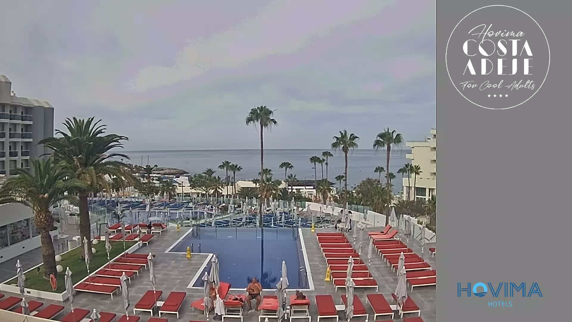 An outdoor pool area with red lounge chairs surrounds a swimming pool, overlooking a calm ocean under an overcast sky.