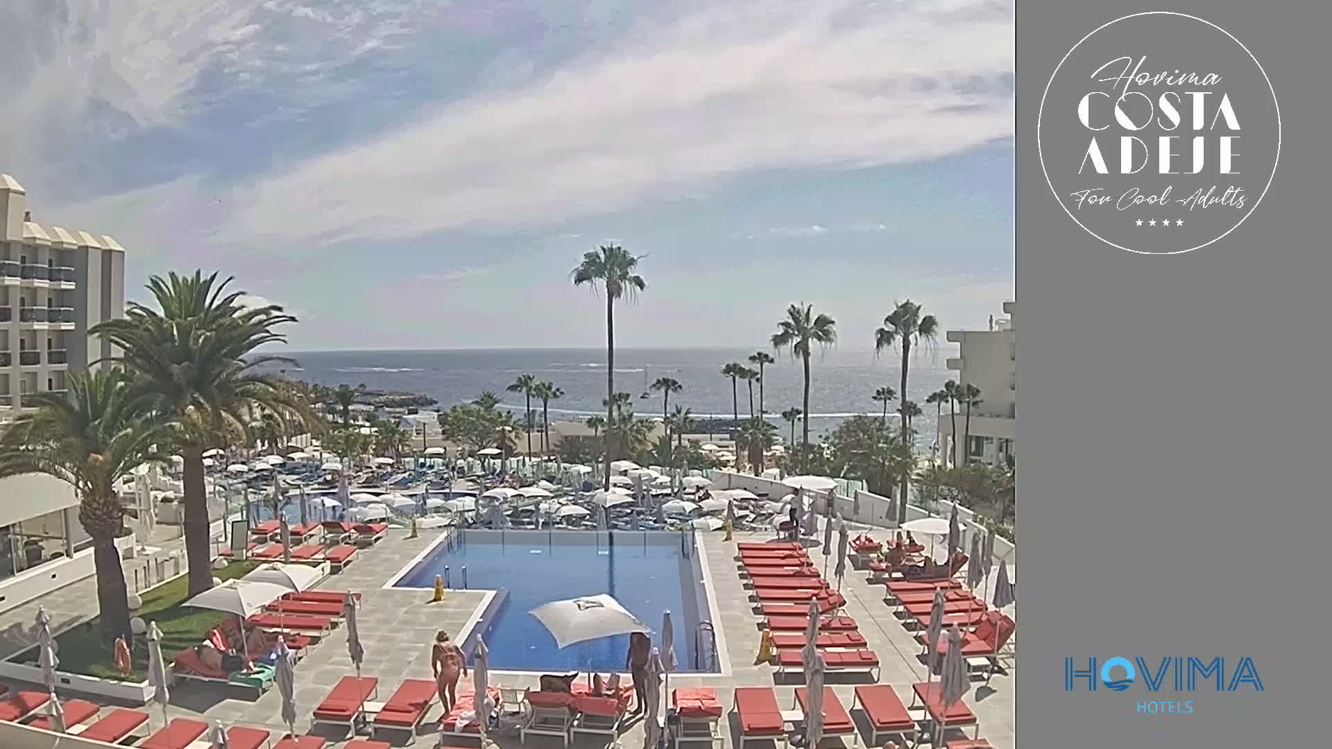A resort pool area with red lounge chairs and umbrellas surrounds a rectangular pool, overlooking a calm ocean under a partly cloudy sky.