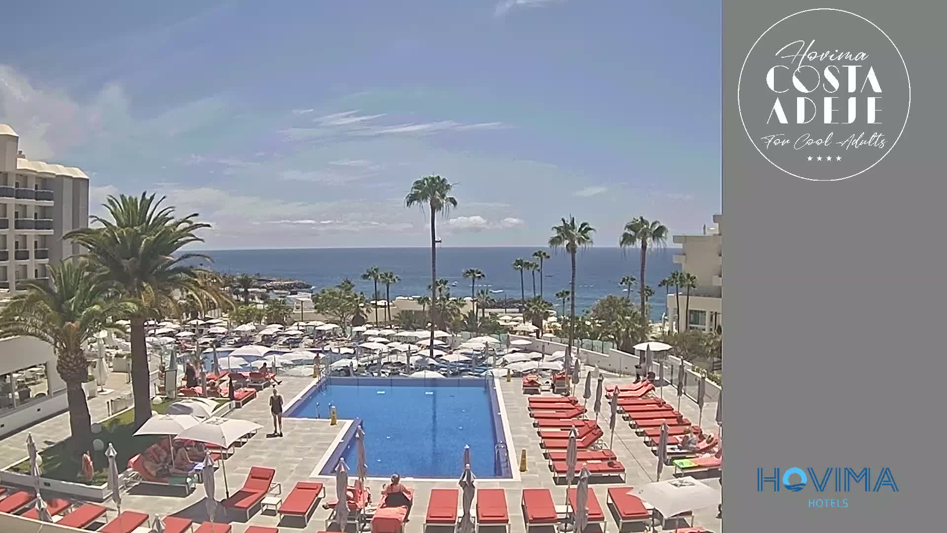 A resort pool area with red lounge chairs and many umbrellas surrounds a large rectangular pool, overlooking a calm ocean under a sunny sky.