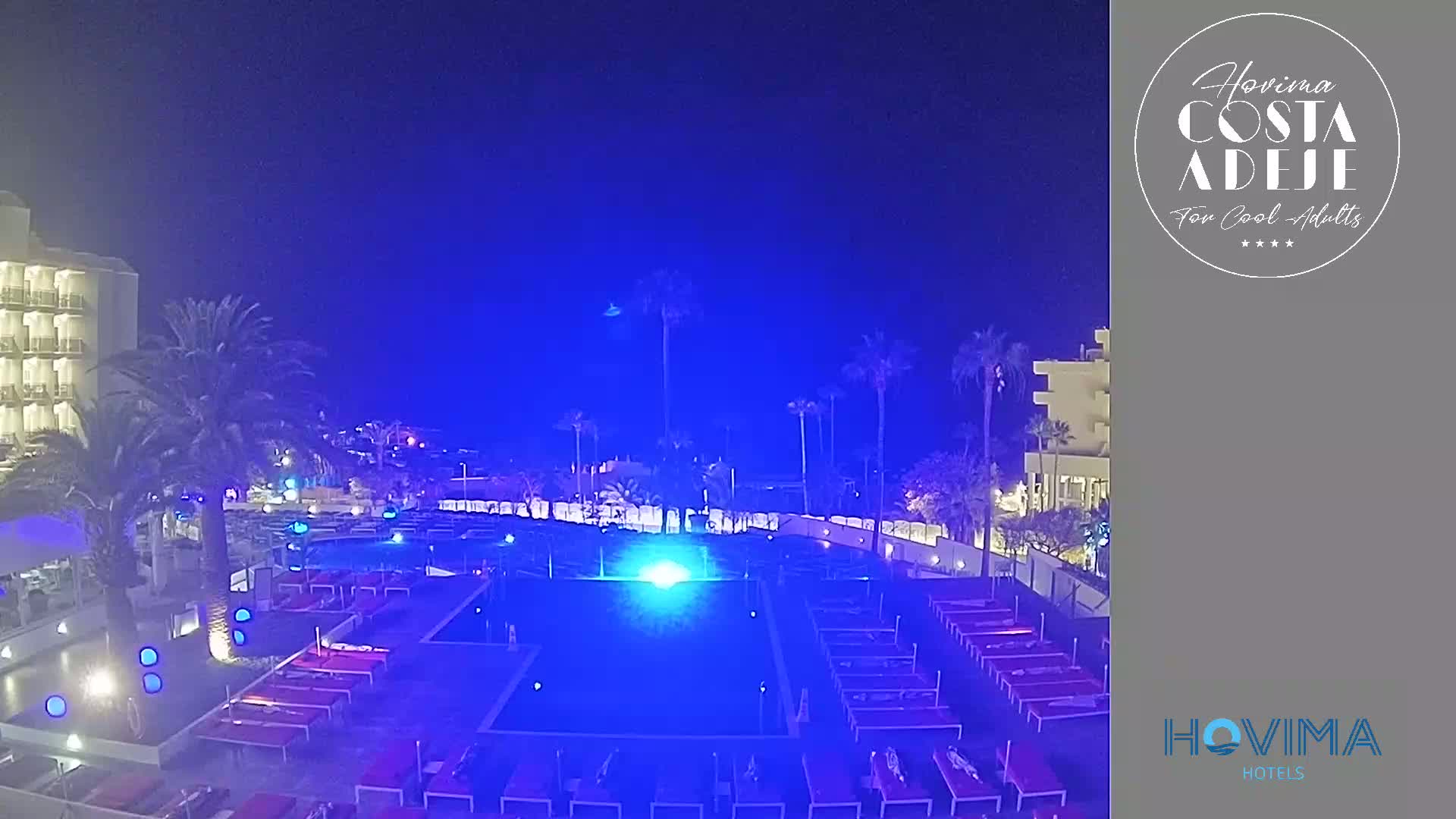 A nighttime view of a resort pool area with palm trees and hotel buildings under a dark blue sky.