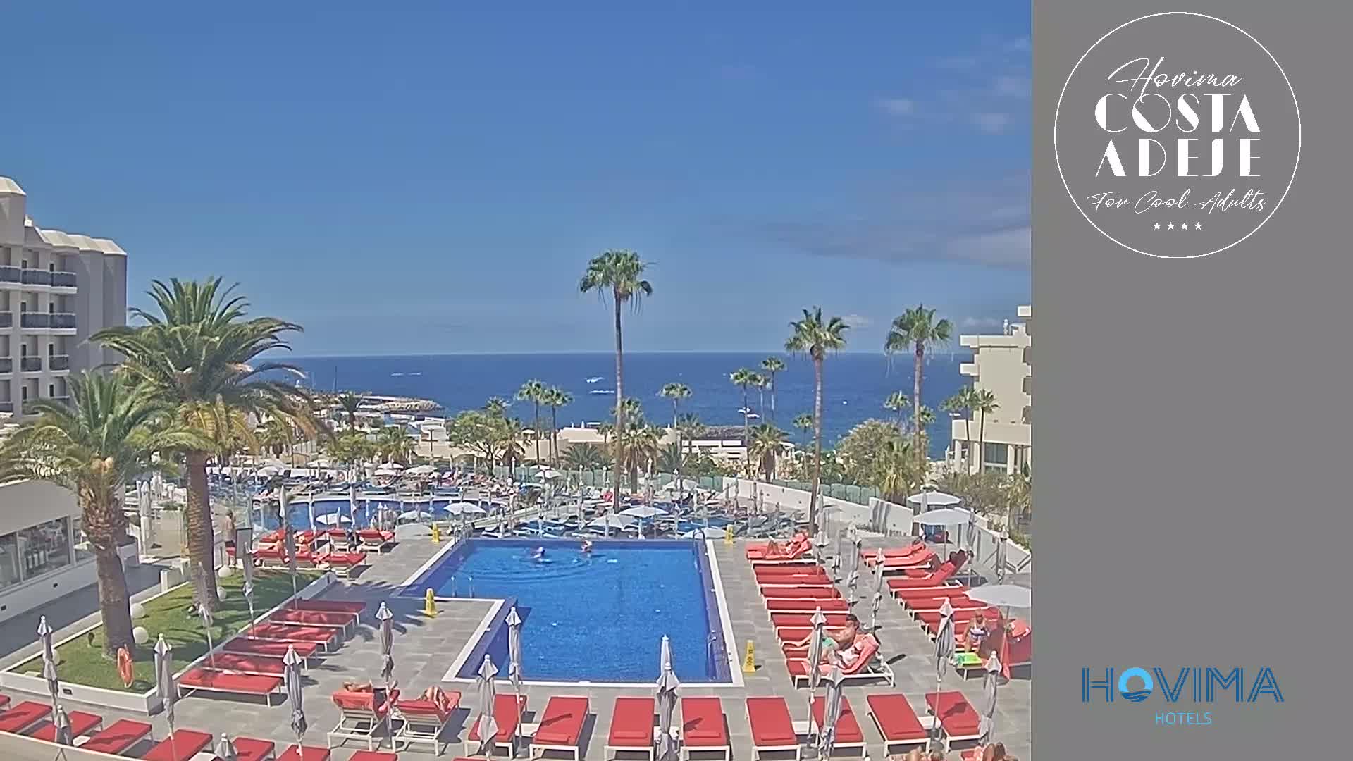 A resort pool area with red lounge chairs and umbrellas overlooks a calm ocean under a clear blue sky.
