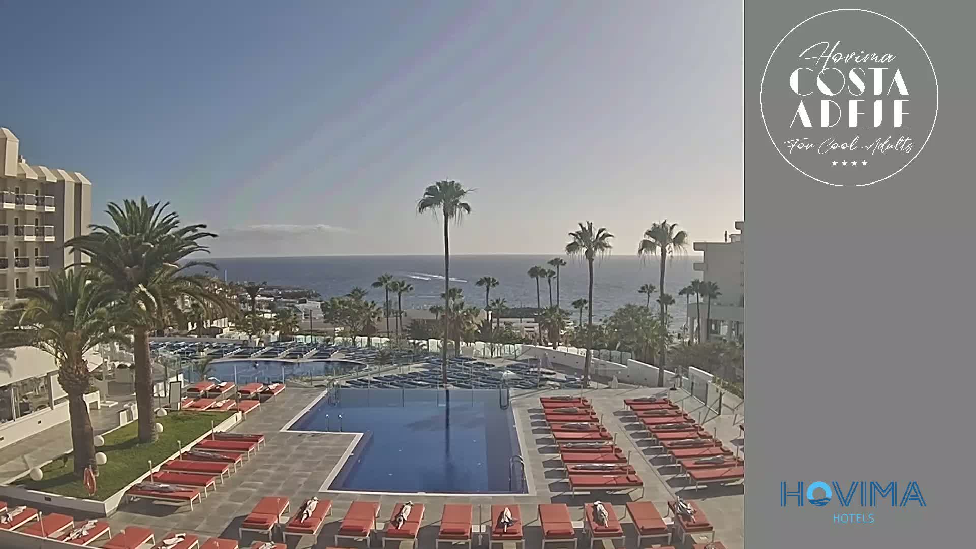A hotel pool area with orange lounge chairs surrounds a swimming pool, overlooking a calm ocean under a clear, sunny sky.