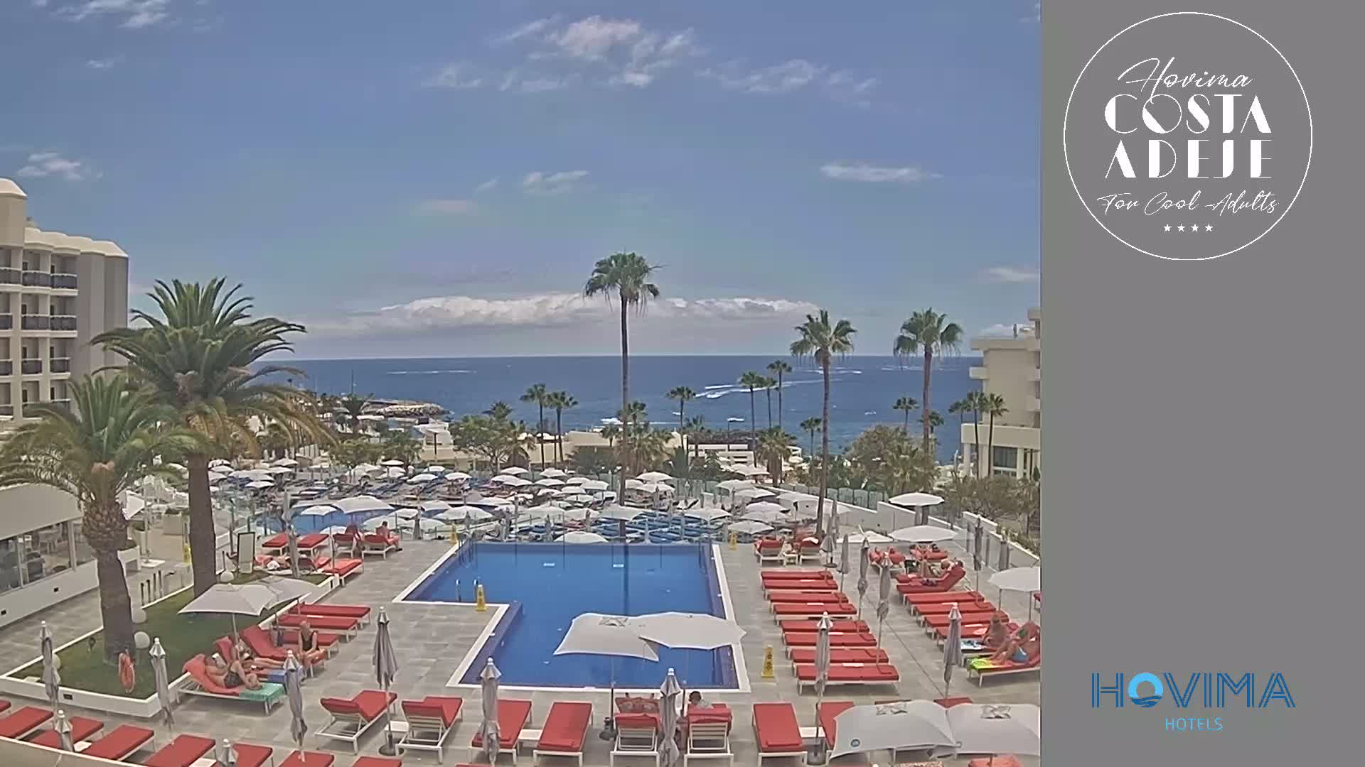 A resort pool area with red lounge chairs and umbrellas surrounds a large rectangular pool, overlooking a calm ocean under a partly cloudy sky.