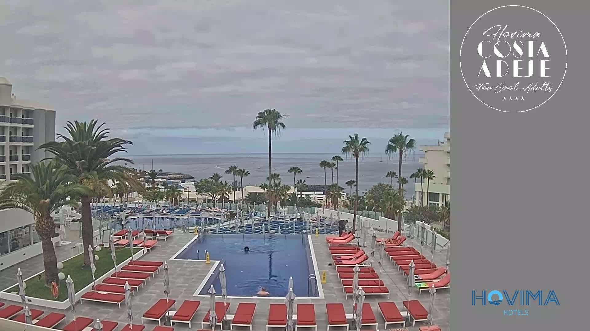 An outdoor pool area with red lounge chairs and umbrellas overlooks a calm ocean under an overcast sky.